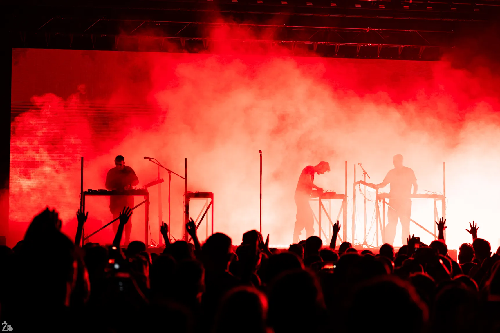 Electronic music performance with three artists on stage and a red smoke-lit background, crowd hands raised.
