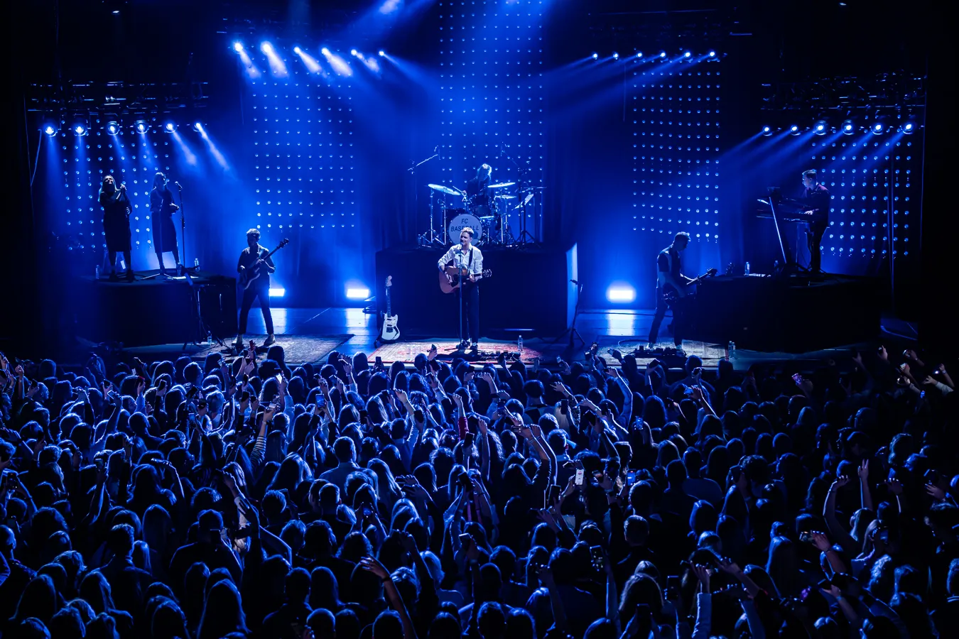 Live band performing on stage with blue lighting in front of a large crowd at a concert.