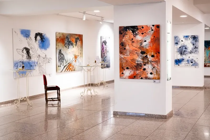 Art gallery room with abstract paintings, a red chair, and small tables with decorative items.