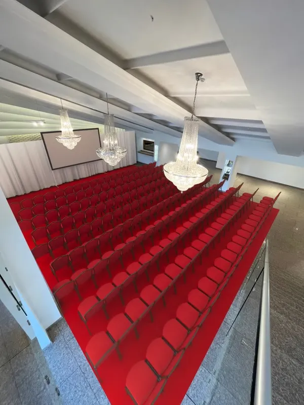Empty auditorium with rows of red chairs facing a projector screen and crystal chandeliers above.