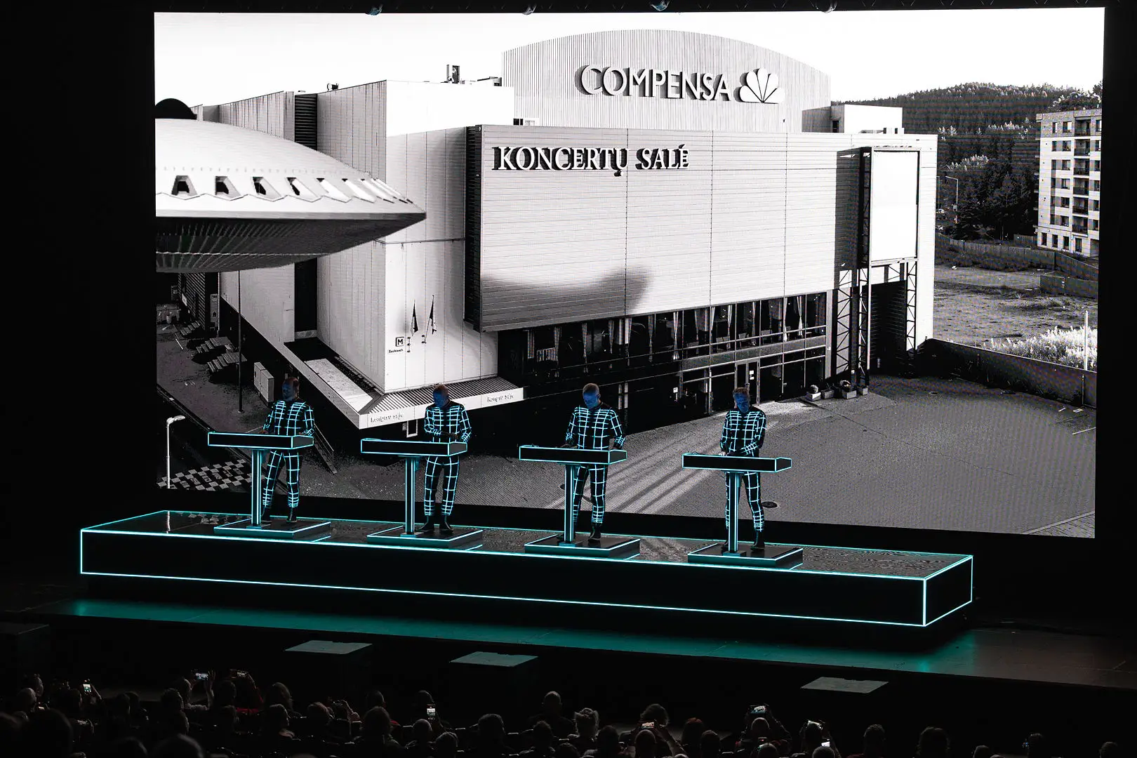 Four performers in illuminated suits stand behind keyboards on a lit stage with a large black-and-white backdrop.