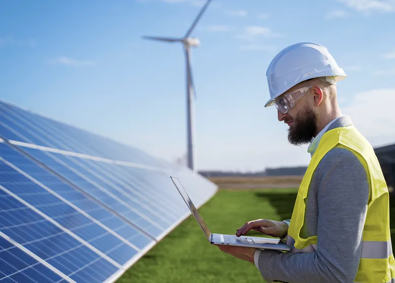 Engineer in safety helmet and vest working on laptop near solar panels with wind turbine in background.