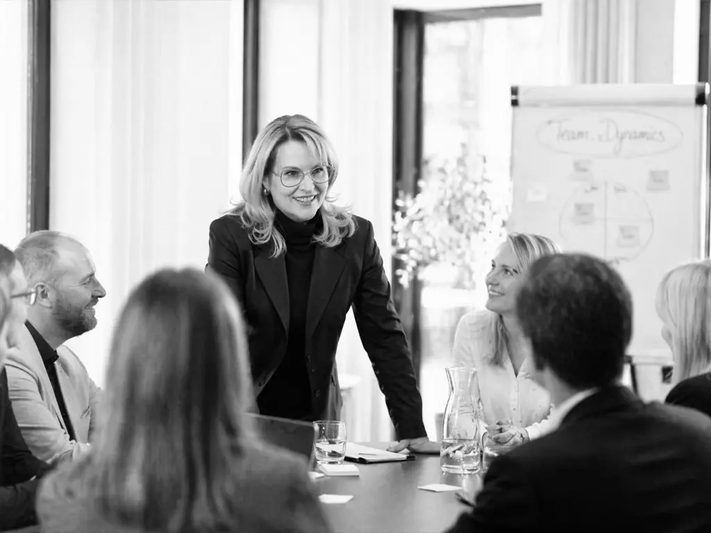 Nicole Flockenhaus moderiert eine Gruppe von Führungskräften an einem Konferenztisch, neben ihr steht ein Flipchart zu Team-Dynamiken.