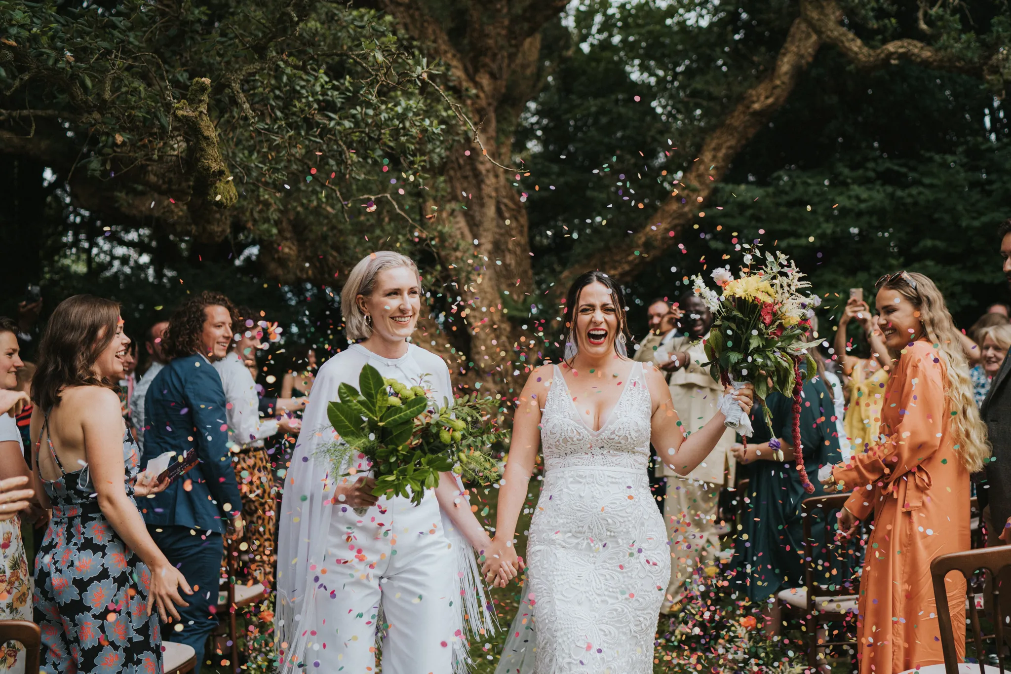 Two brides holding hands and smiling while walking through confetti at an outdoor wedding ceremony.