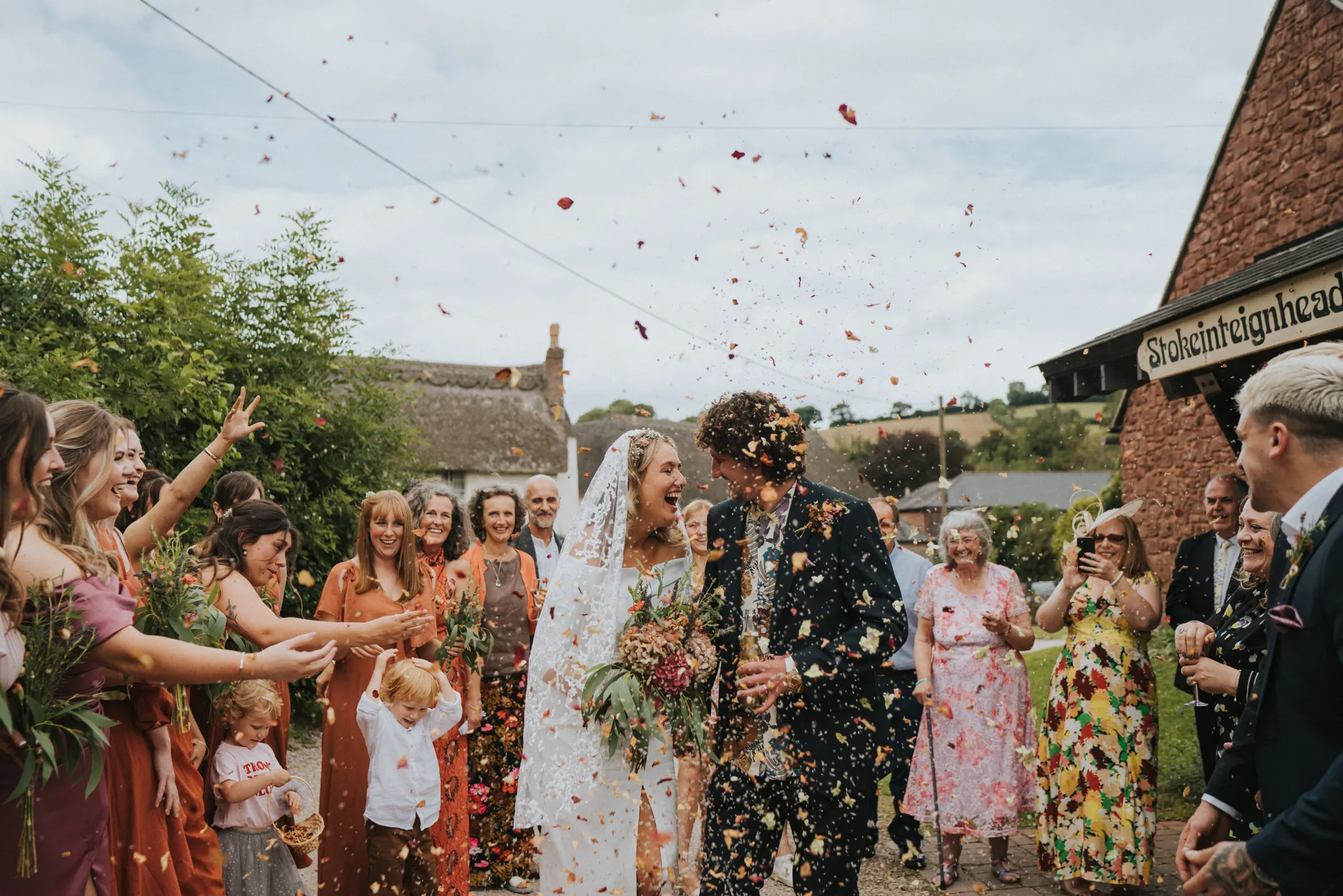 Bride and groom laughing while guests throw flower petals at their outdoor wedding celebration.