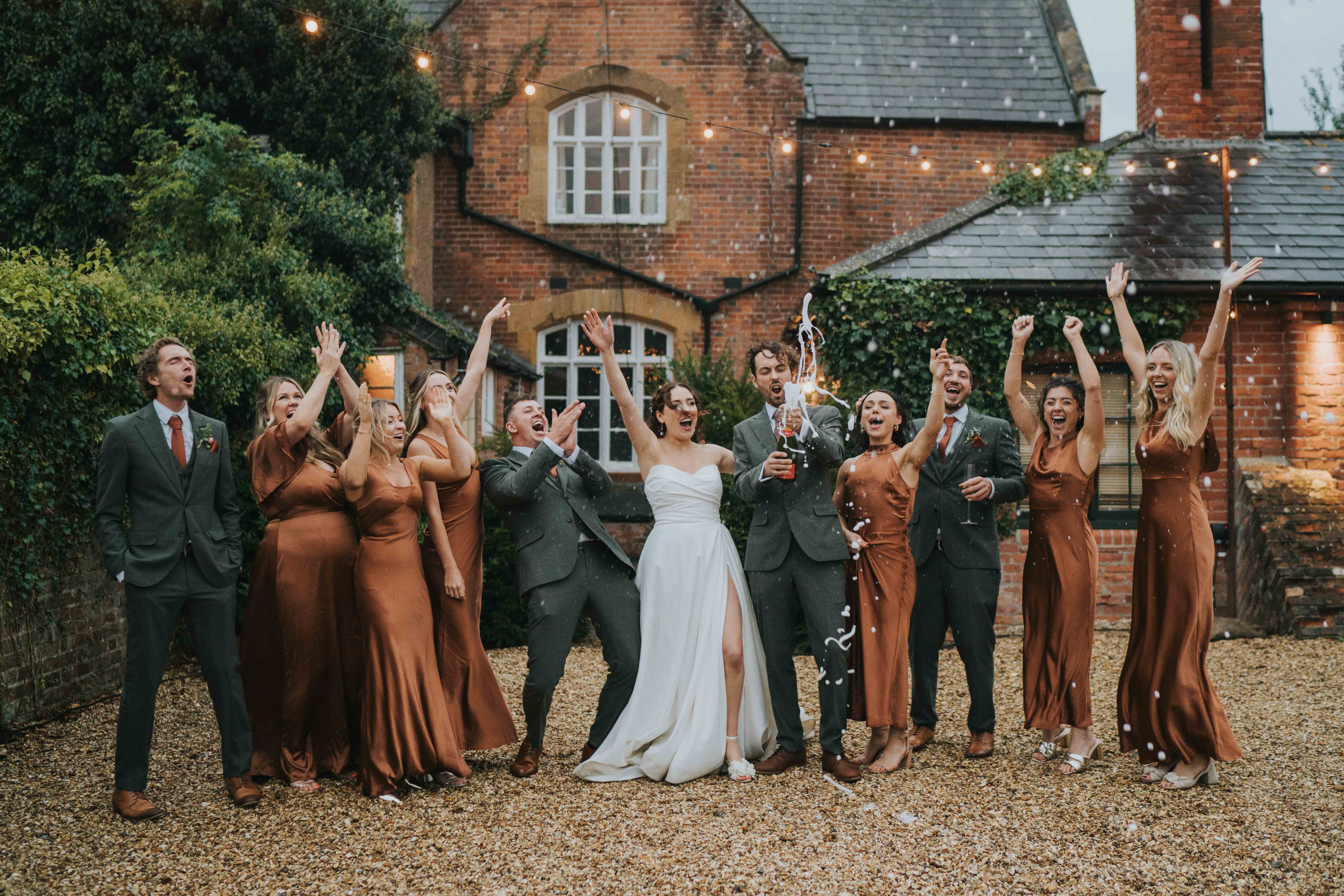 Wedding party outdoors in front of a brick house celebrating with champagne and raised arms.
