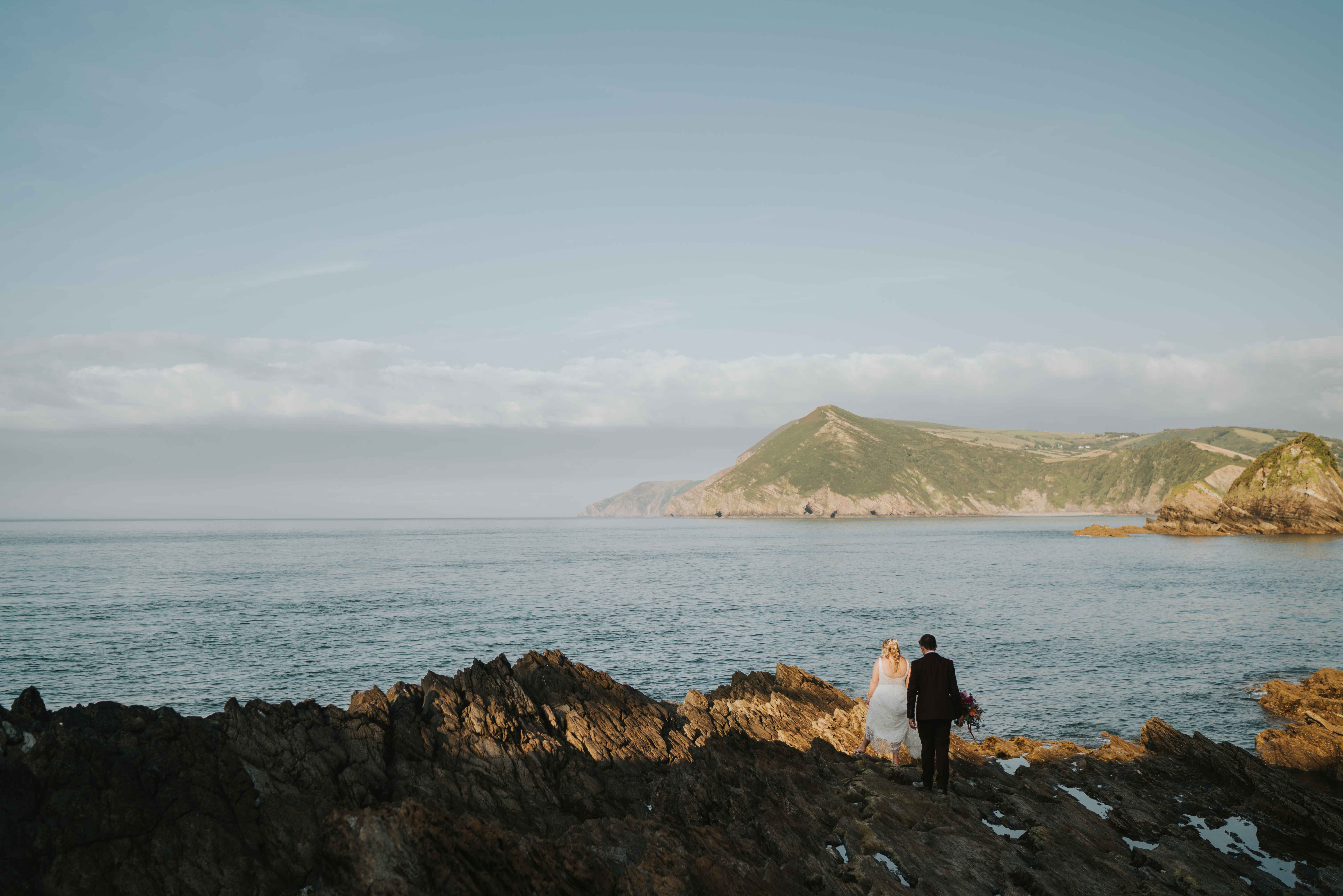 Bride and groom walking hand in hand on rocky shore with calm sea and green hills in the background.