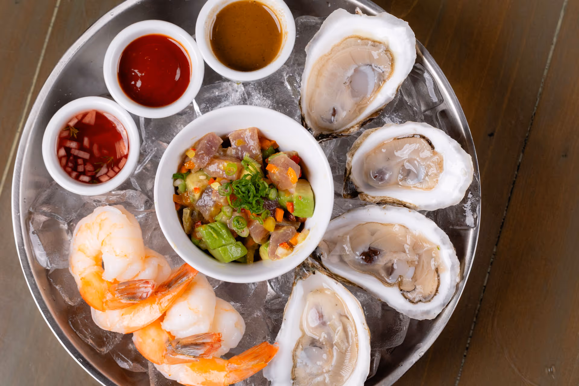 Metal platter with oysters, shrimp, diced seafood and avocado salad, and three dipping sauces on ice.