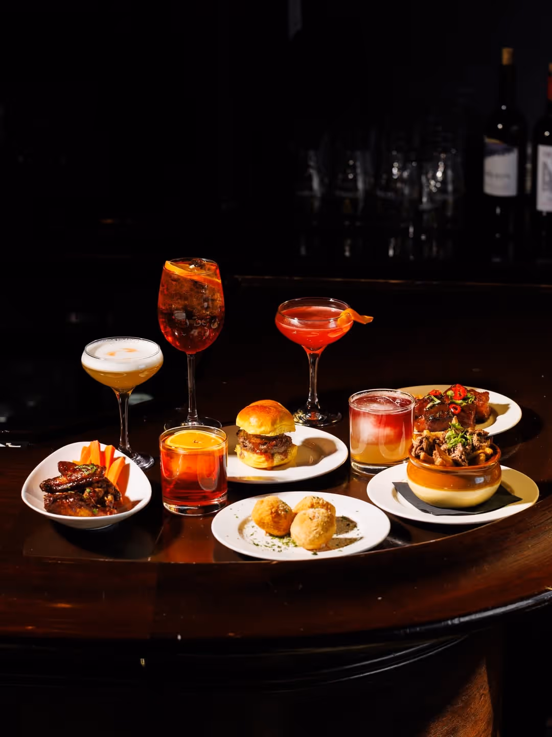Varied cocktails and appetizer plates including wings, sliders, croquettes, and small dishes on a dark table.