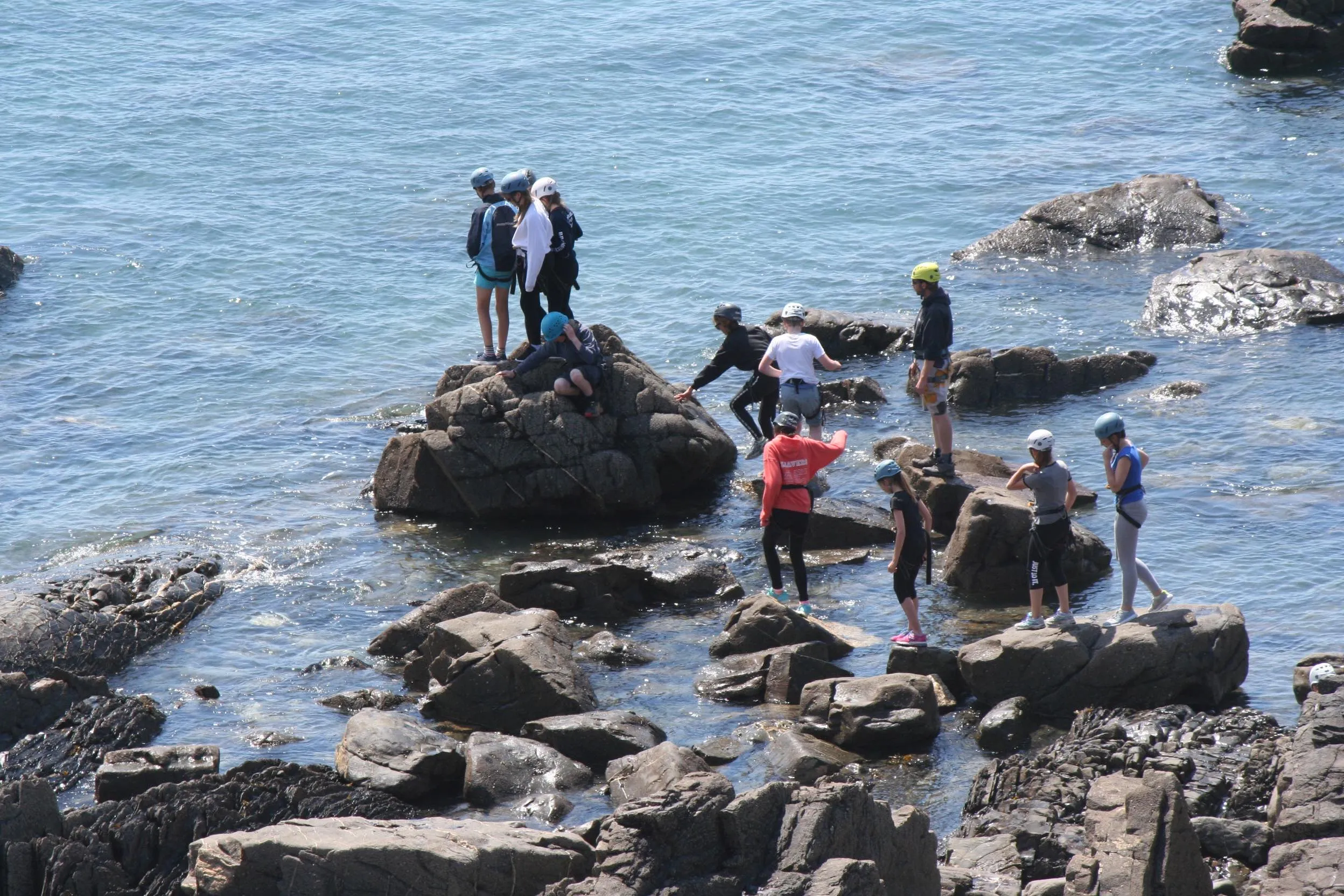 Group of people wearing helmets and outdoor gear climbing and standing on rocky outcrops by the sea.