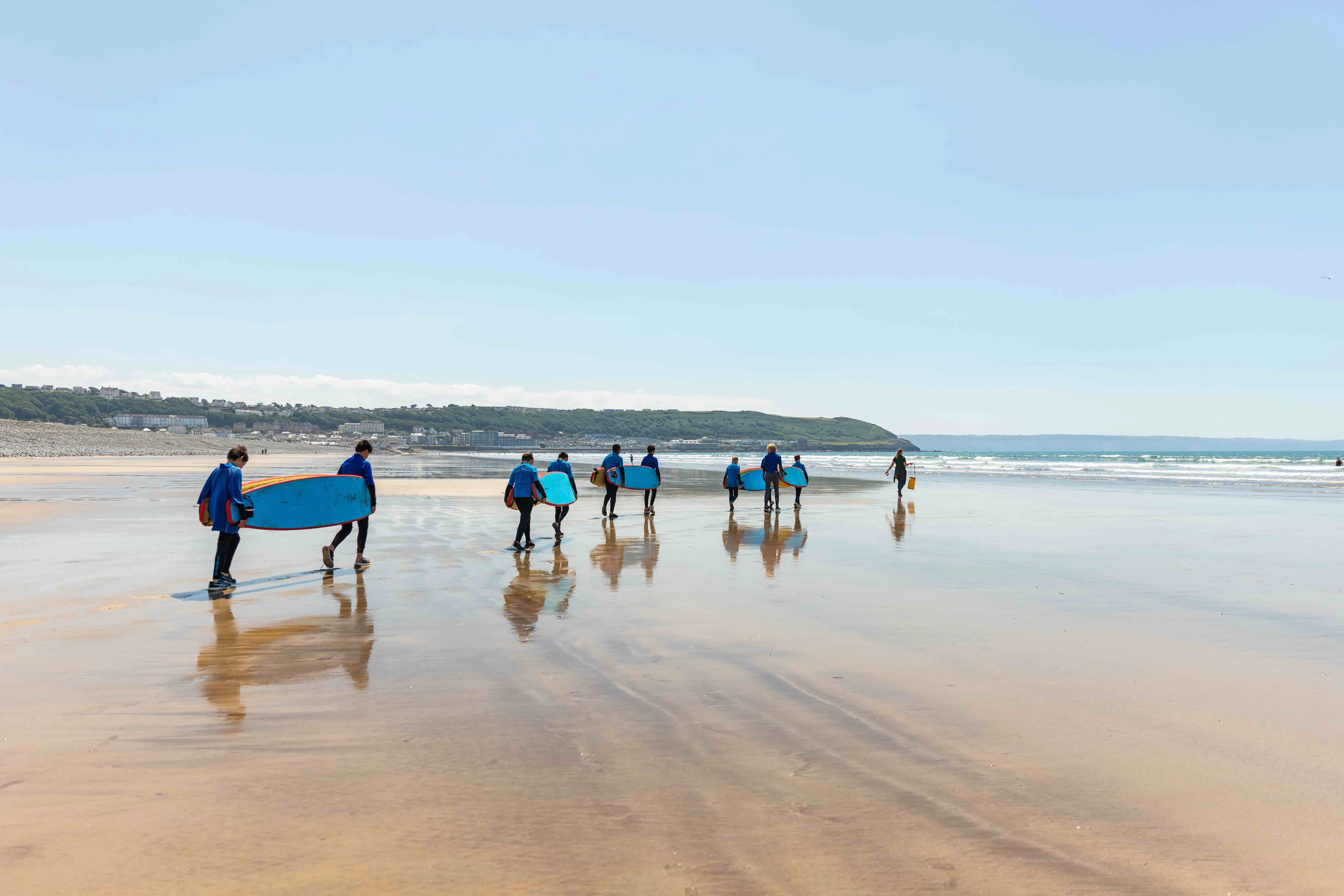 Group of children in wetsuits carrying surfboards walking towards the sea on a sandy beach under a clear blue sky.