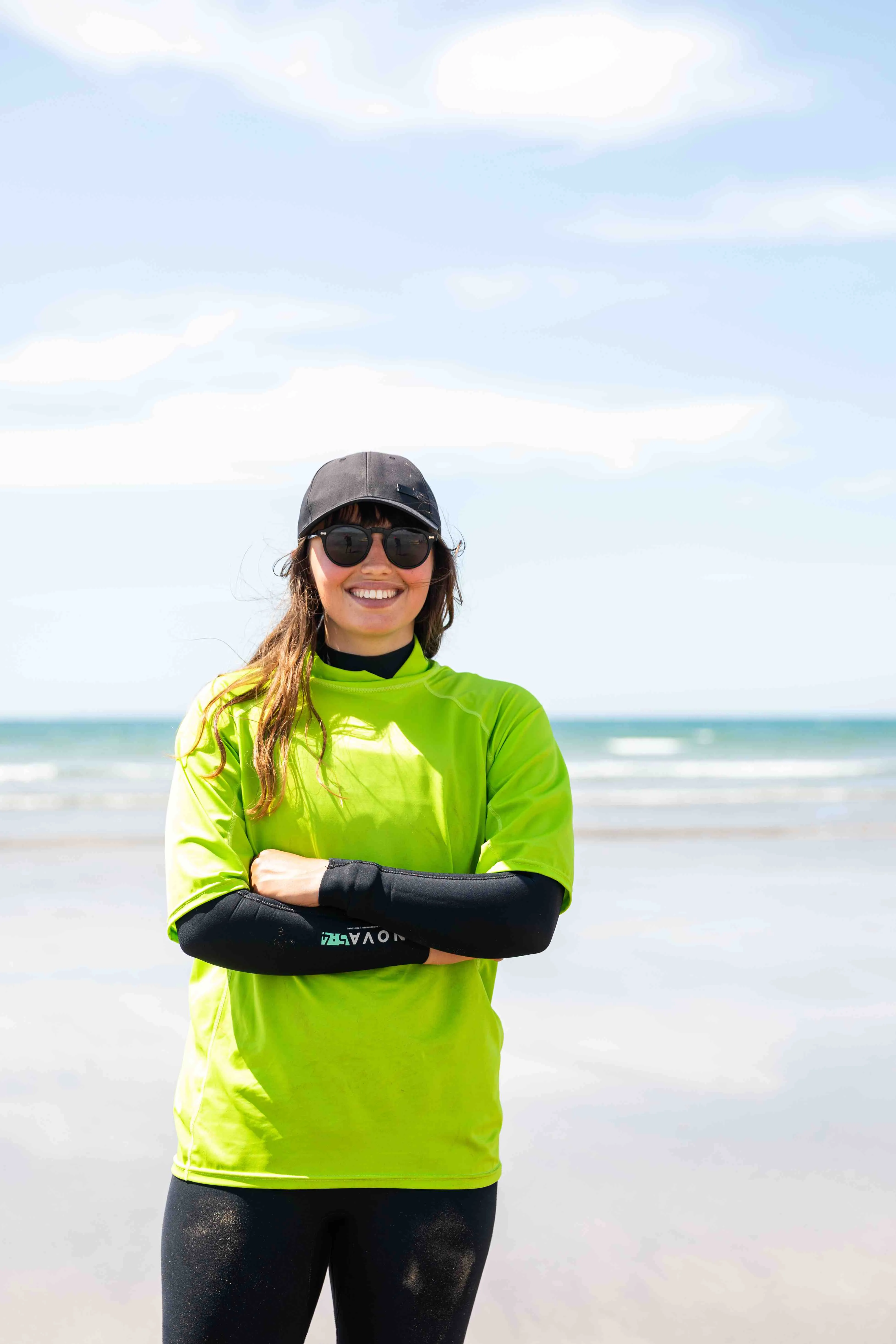 Smiling woman wearing sunglasses, a black cap, and a bright green shirt standing with arms crossed on a beach.