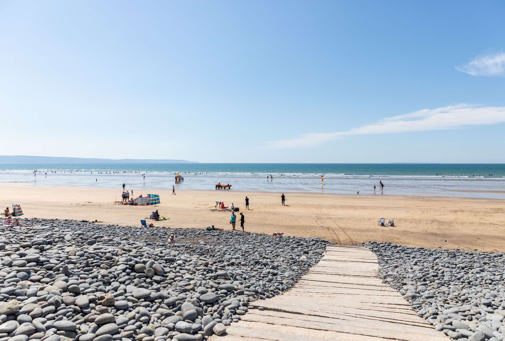 Wooden boardwalk leading over a pebble beach to a sandy shore with people enjoying a clear sunny day by the sea.