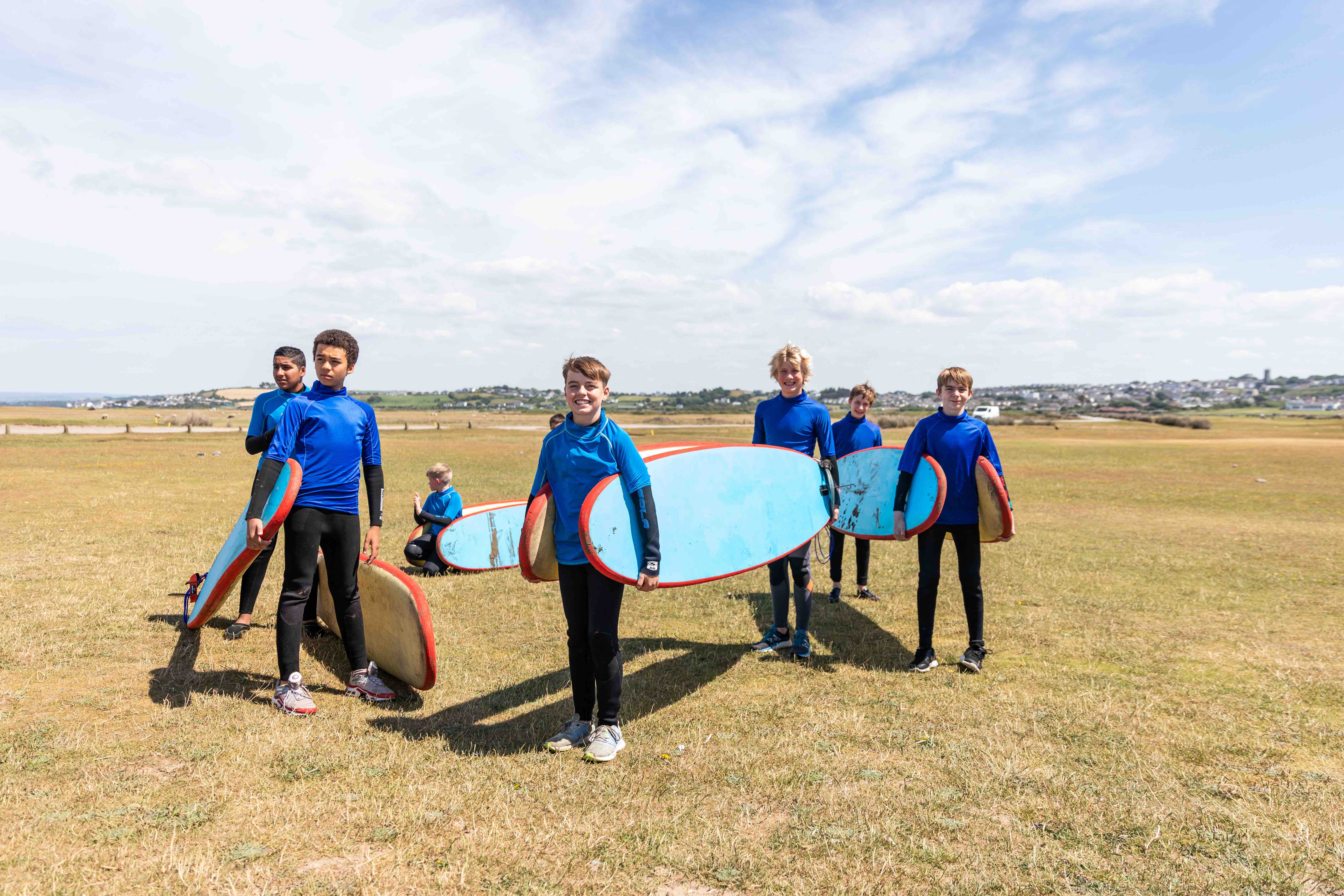 Group of boys in blue wetsuits standing on grass holding blue surfboards with red edges under a partly cloudy sky.
