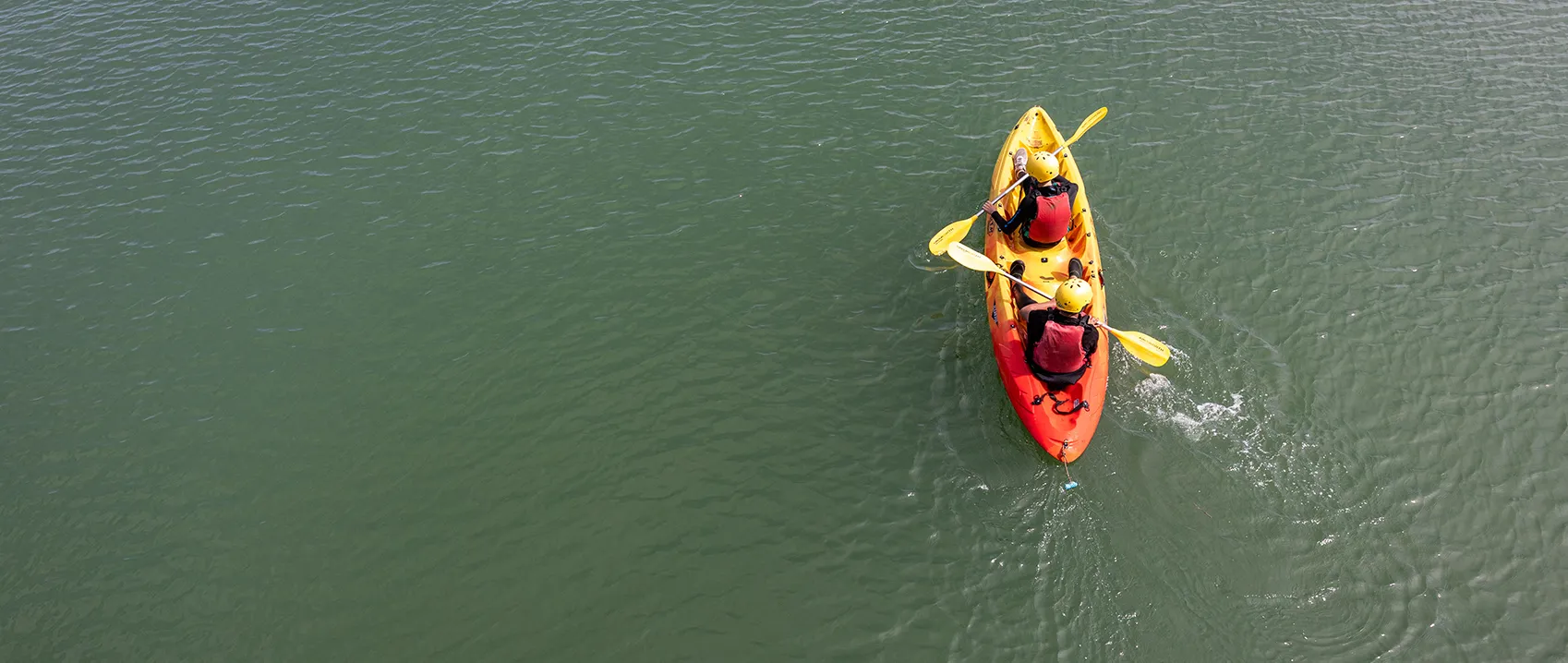 Two people wearing yellow helmets and red life jackets paddling a yellow and red kayak on calm green water.