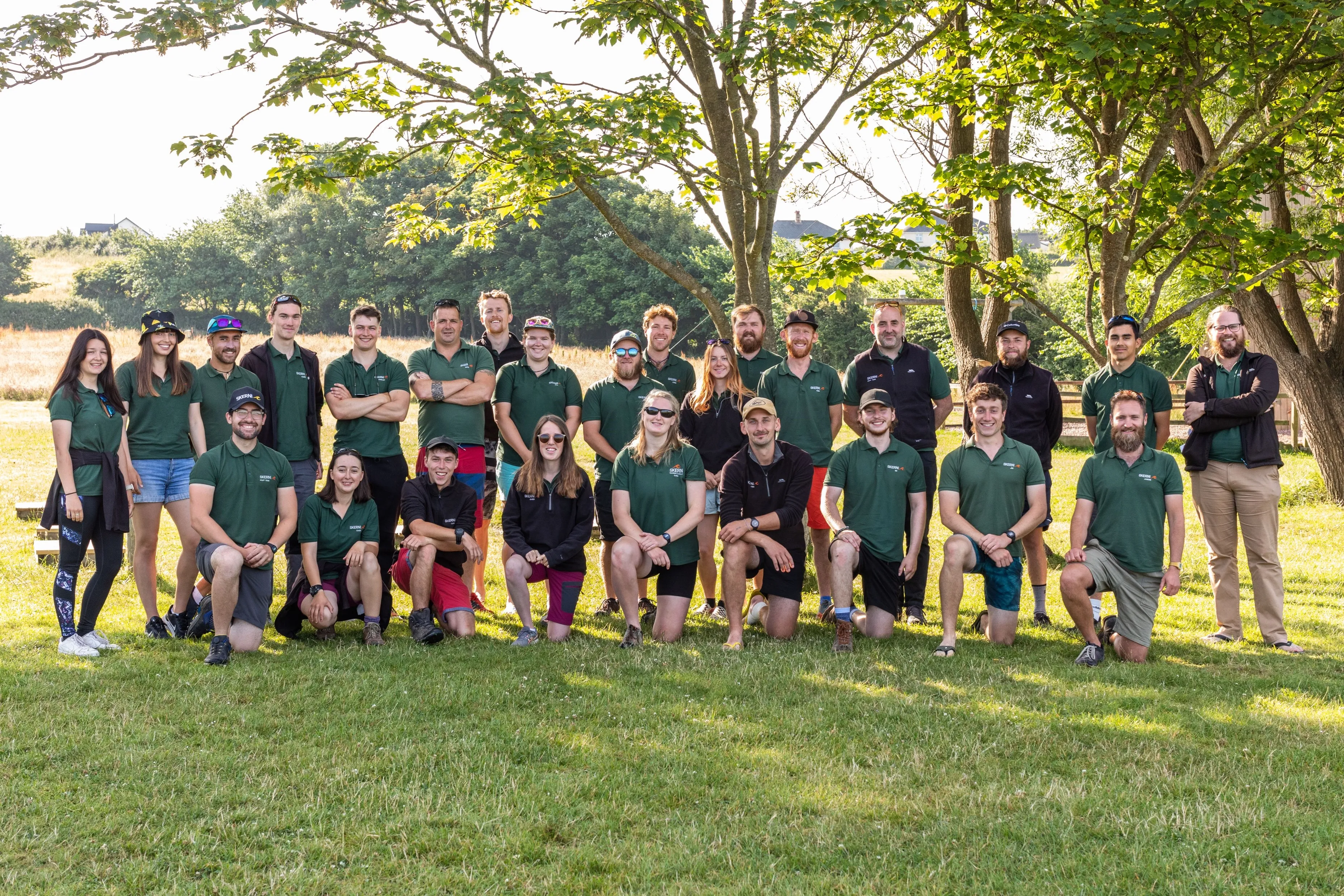 Group of 26 people posing outdoors on grass with trees and countryside in the background, many wearing green polo shirts.