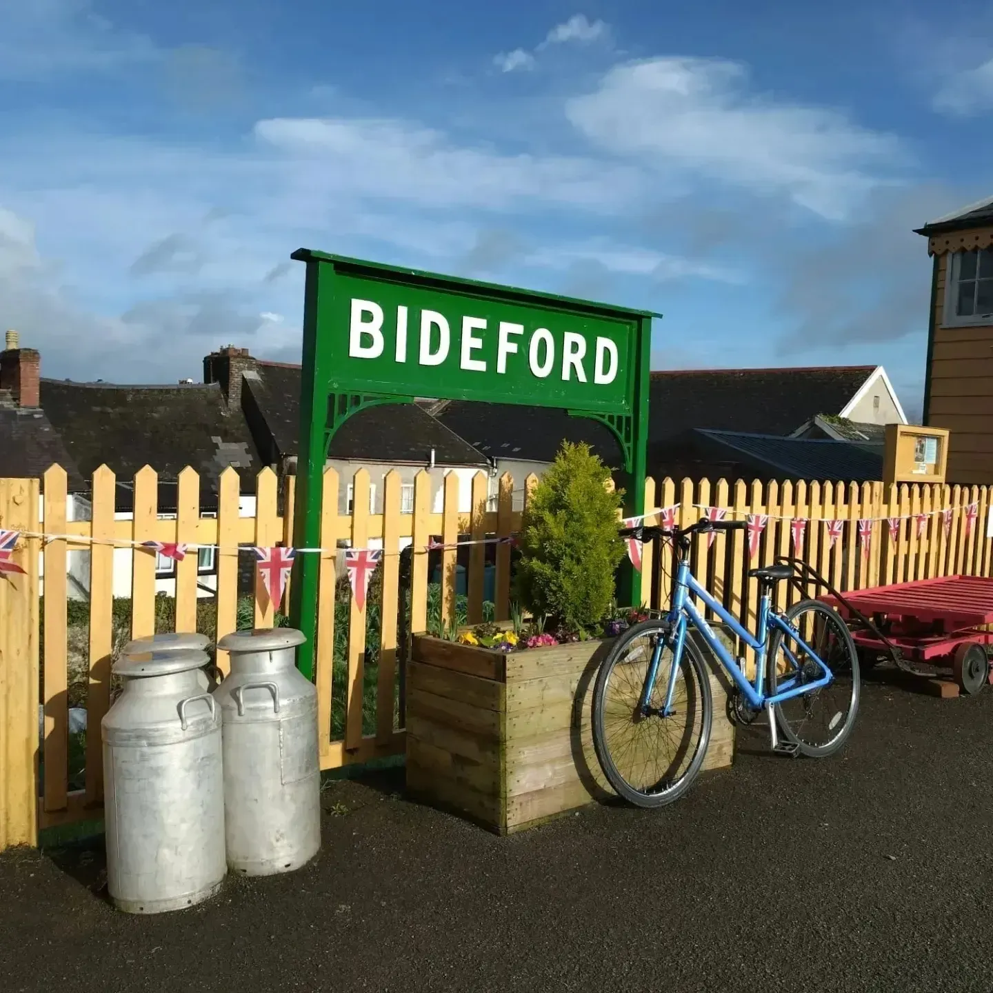 Green Bideford sign at a train station platform with a yellow picket fence, two metal milk churns, a blue bicycle, and a wooden planter with flowers.