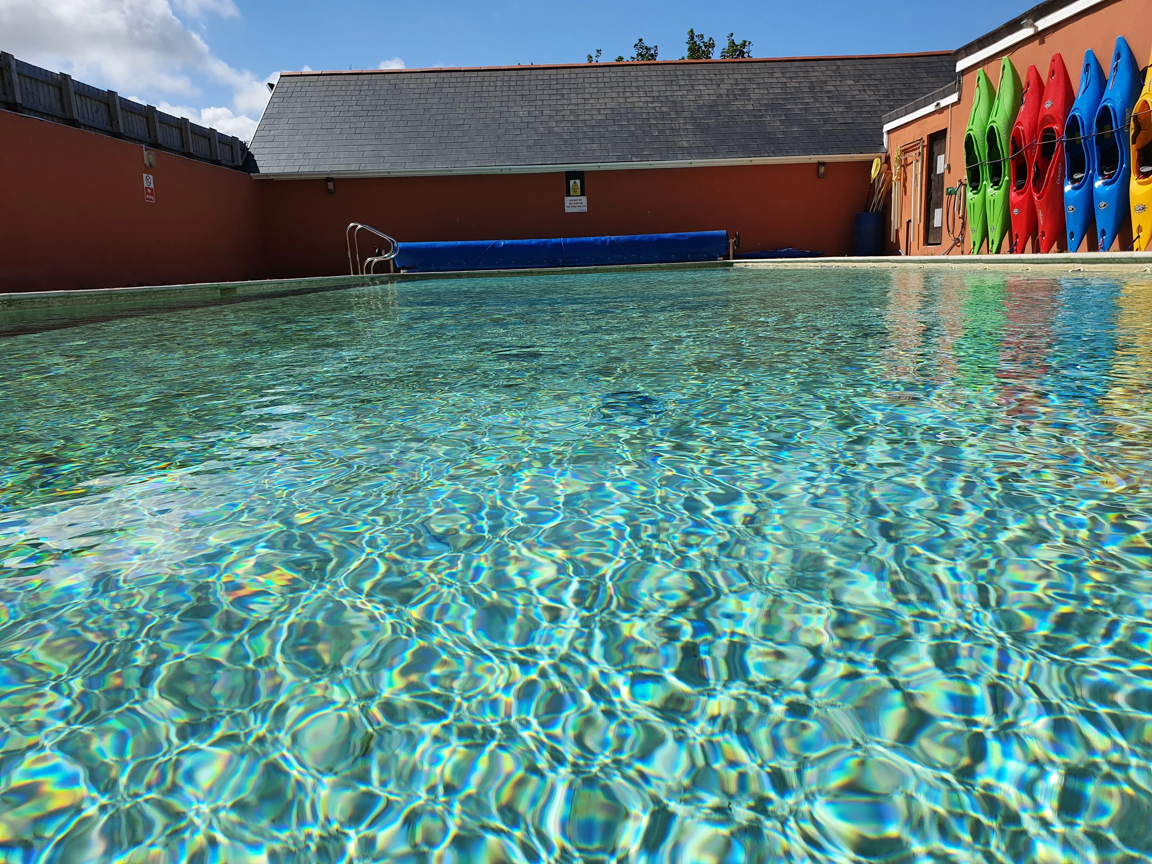 Clear outdoor swimming pool with a row of colourful kayaks lined up against an orange wall on the right side.