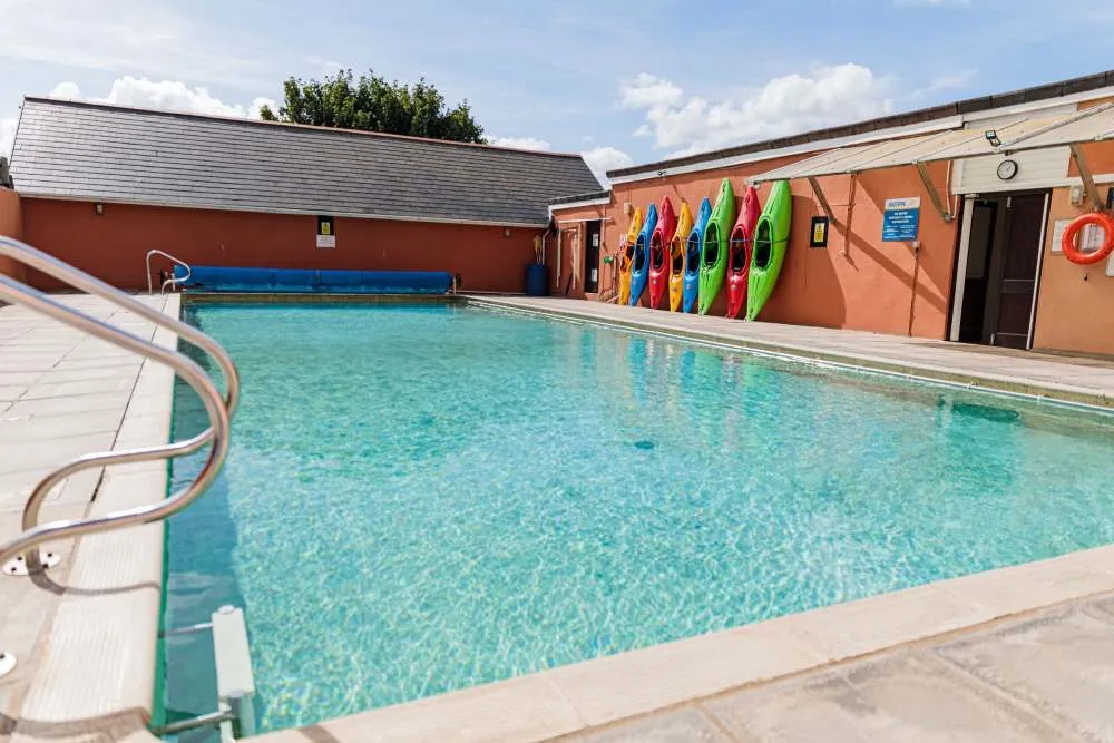 Outdoor swimming pool with clear blue water next to a building with colourful kayaks lined up against the wall.
