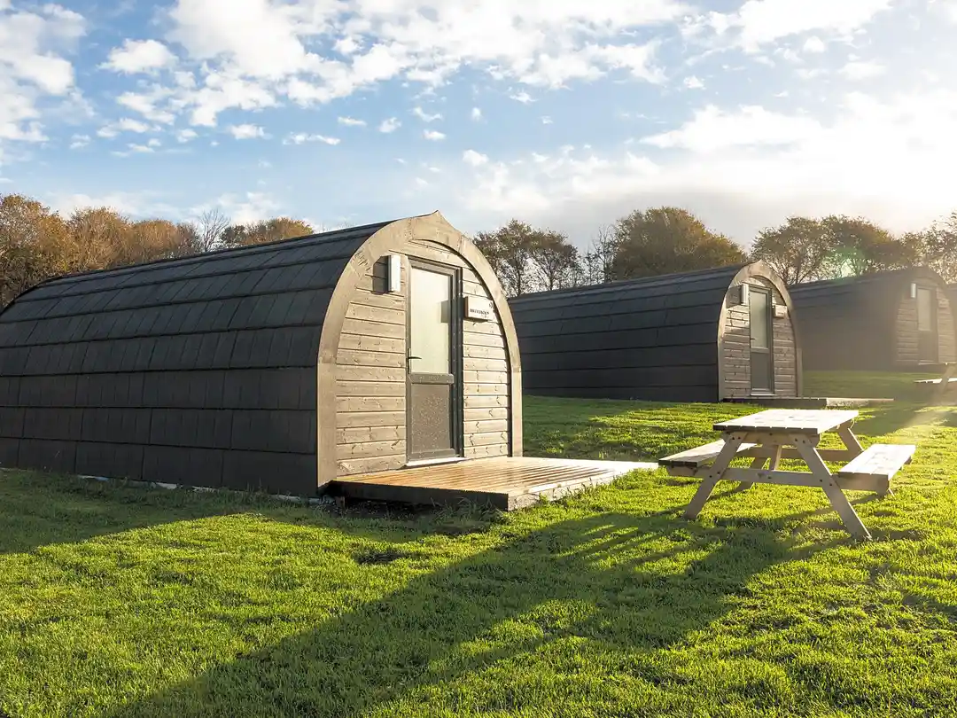 Three black wooden camping pods on green grass under a blue sky with clouds, with a wooden picnic bench in the foreground.