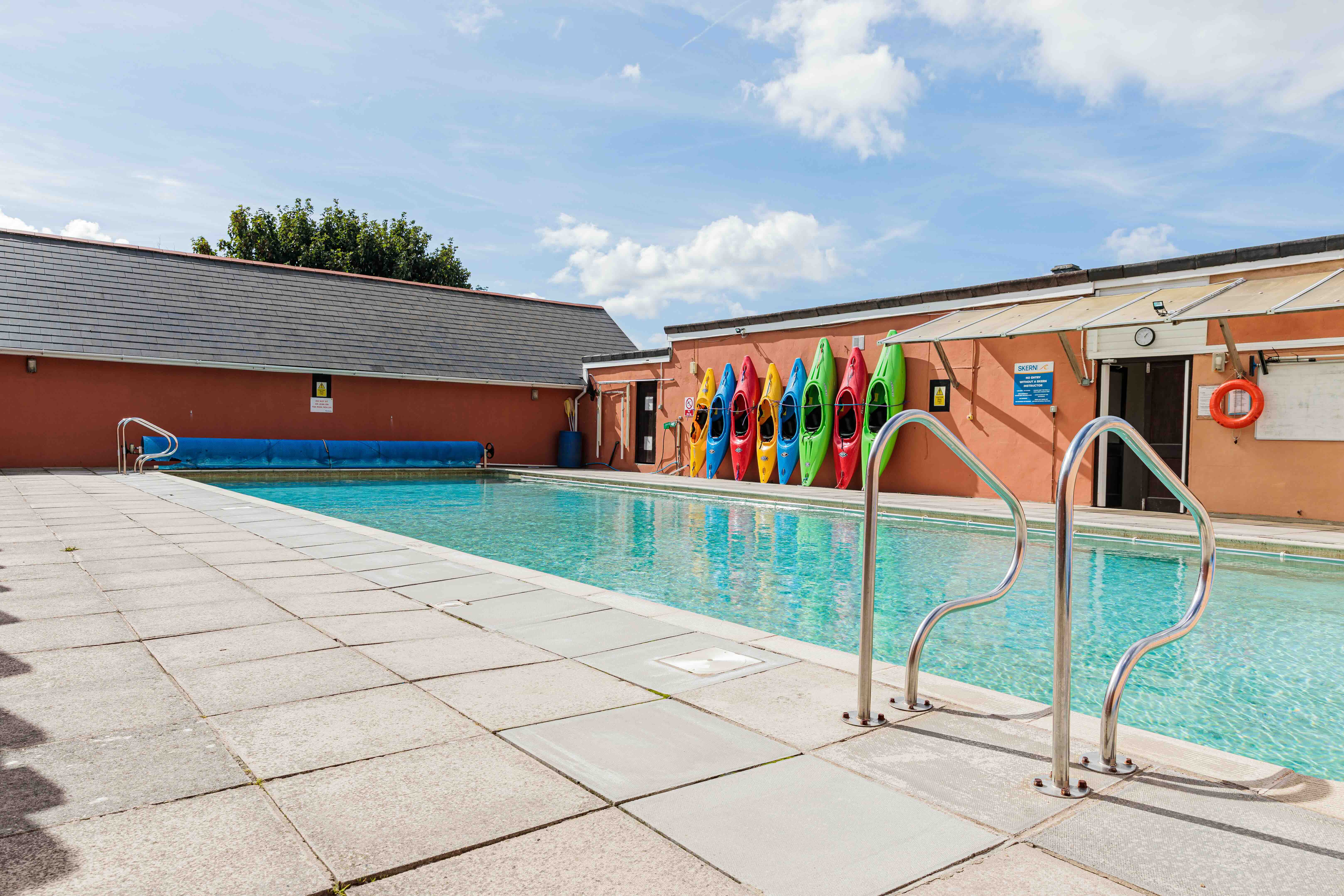 Outdoor swimming pool with colourfully painted kayaks hanging on a wall and metal handrails under a partly cloudy sky.