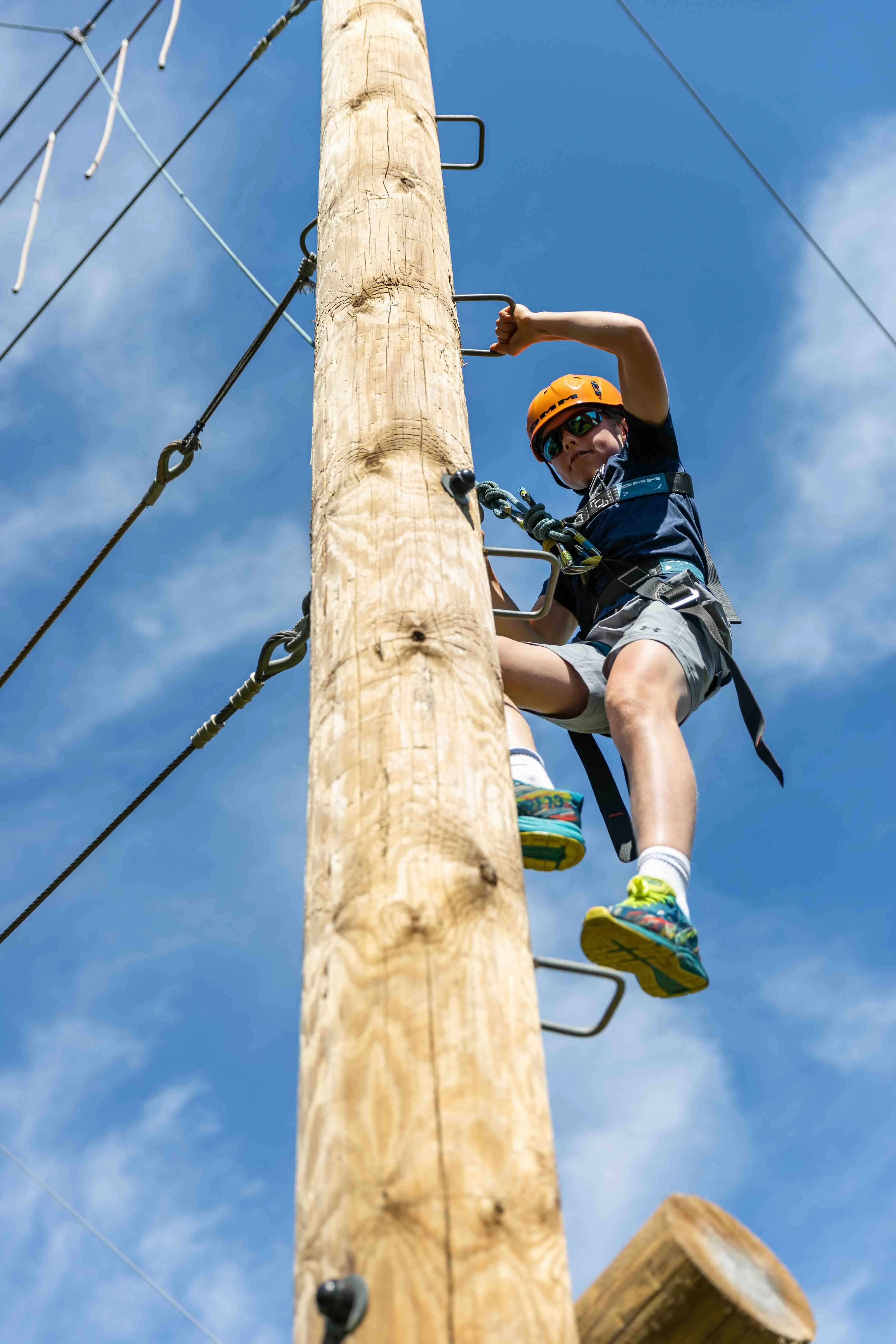 Child wearing helmet and harness climbing a wooden pole with metal rungs against a blue sky.