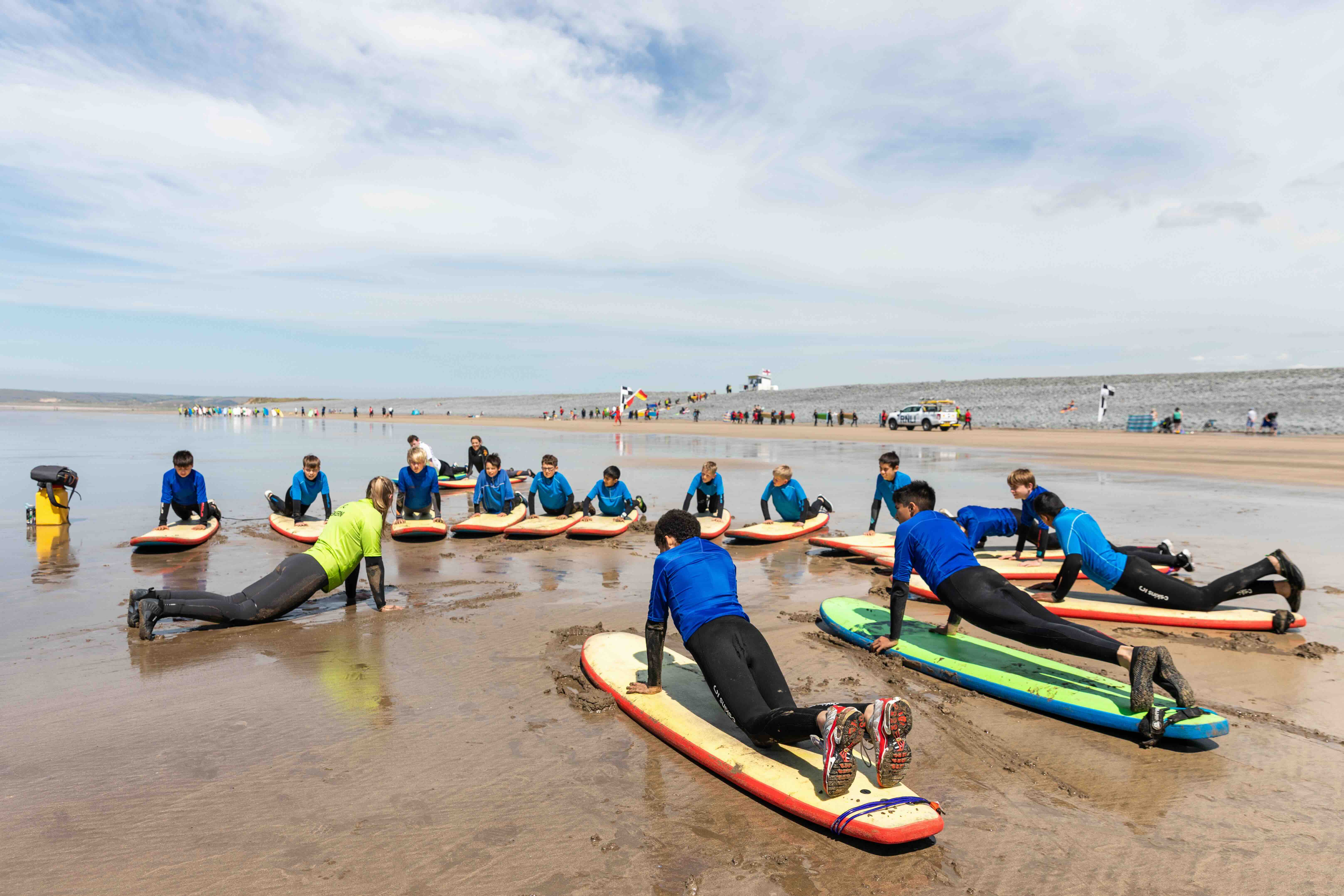 Group of children in wetsuits lying on surfboards on the beach practicing paddling techniques with an instructor in a green jacket.