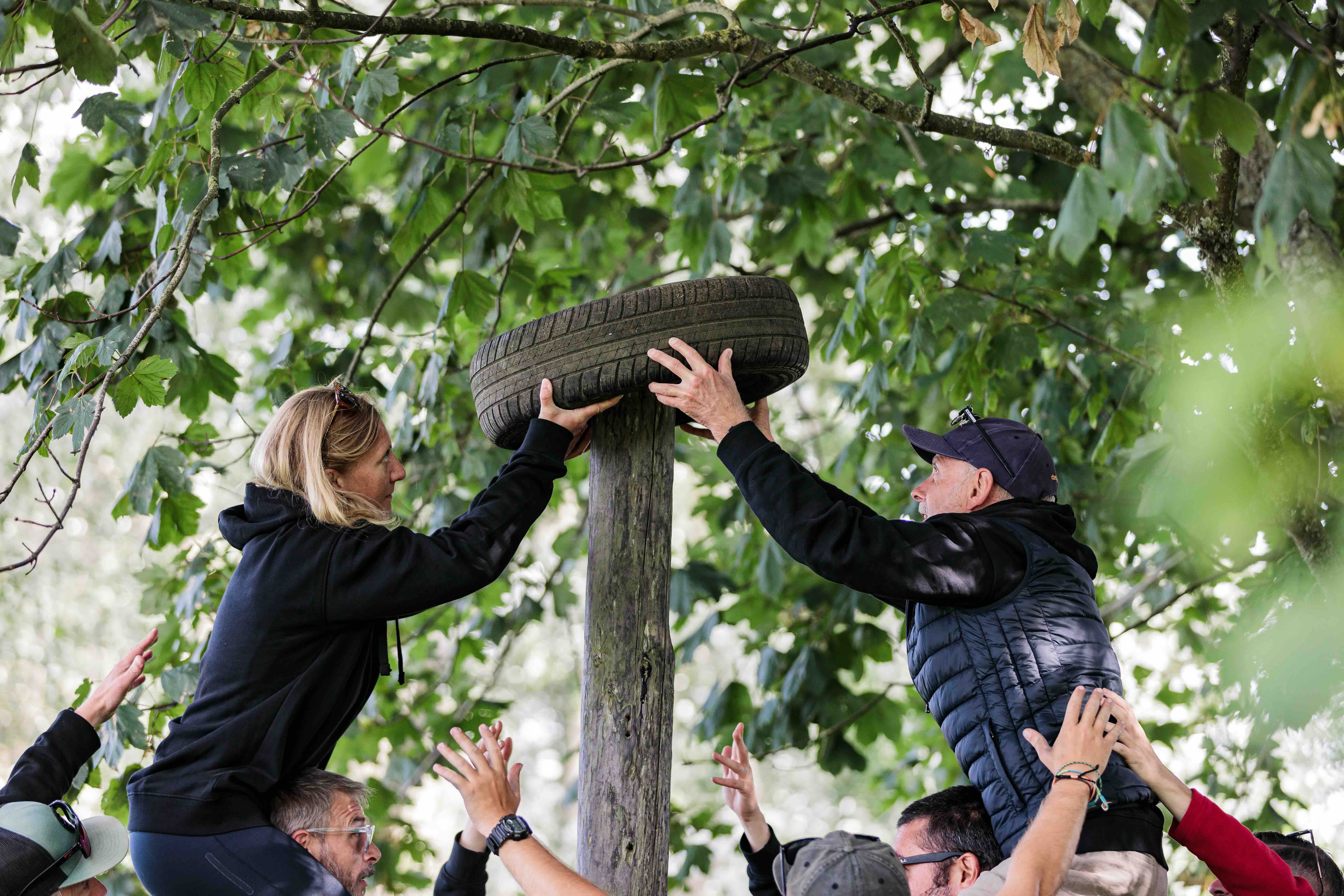Two people supported by others reaching to place a tyre on top of a wooden post under leafy trees.