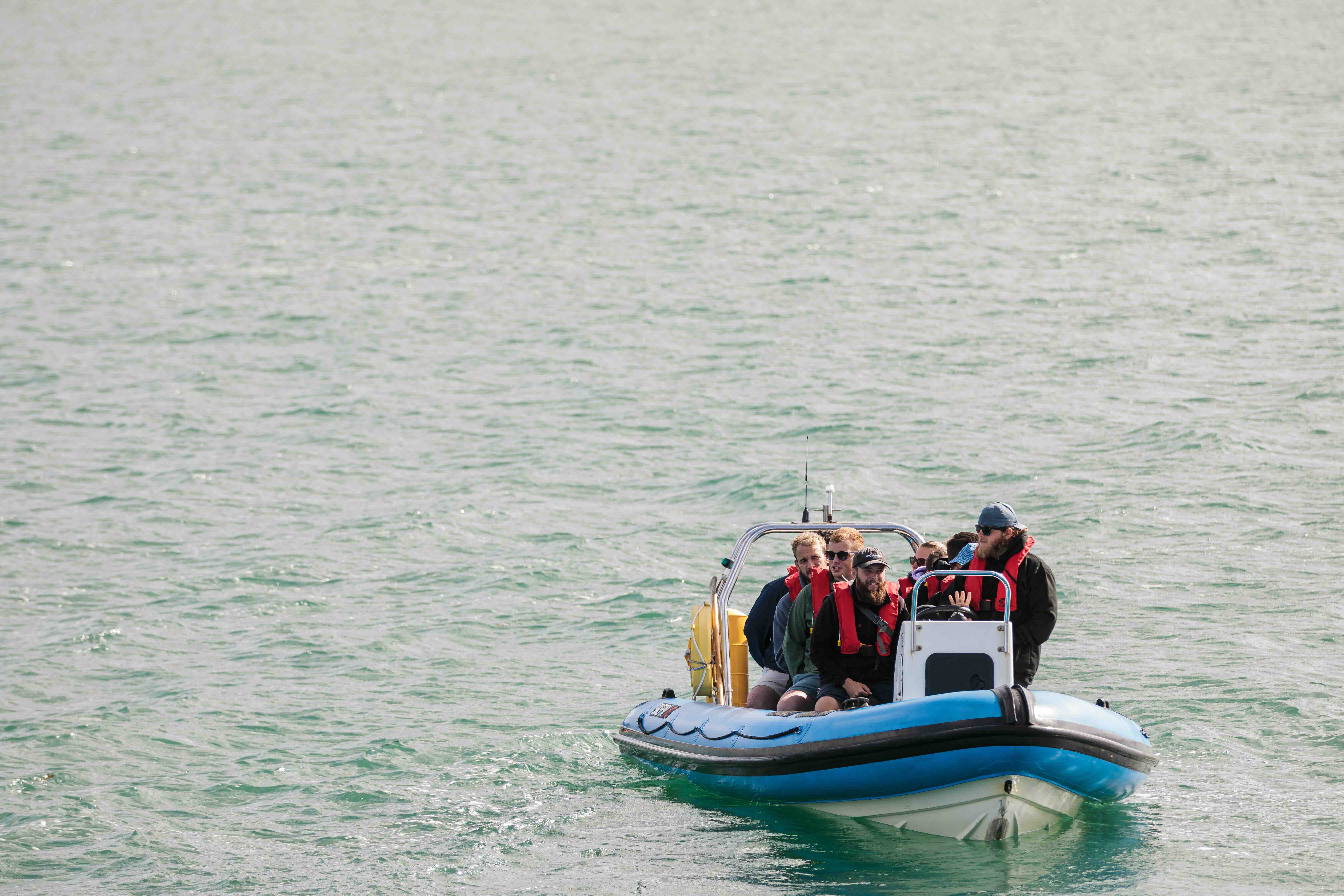 Six people wearing red life jackets on a small blue motorboat in calm water.