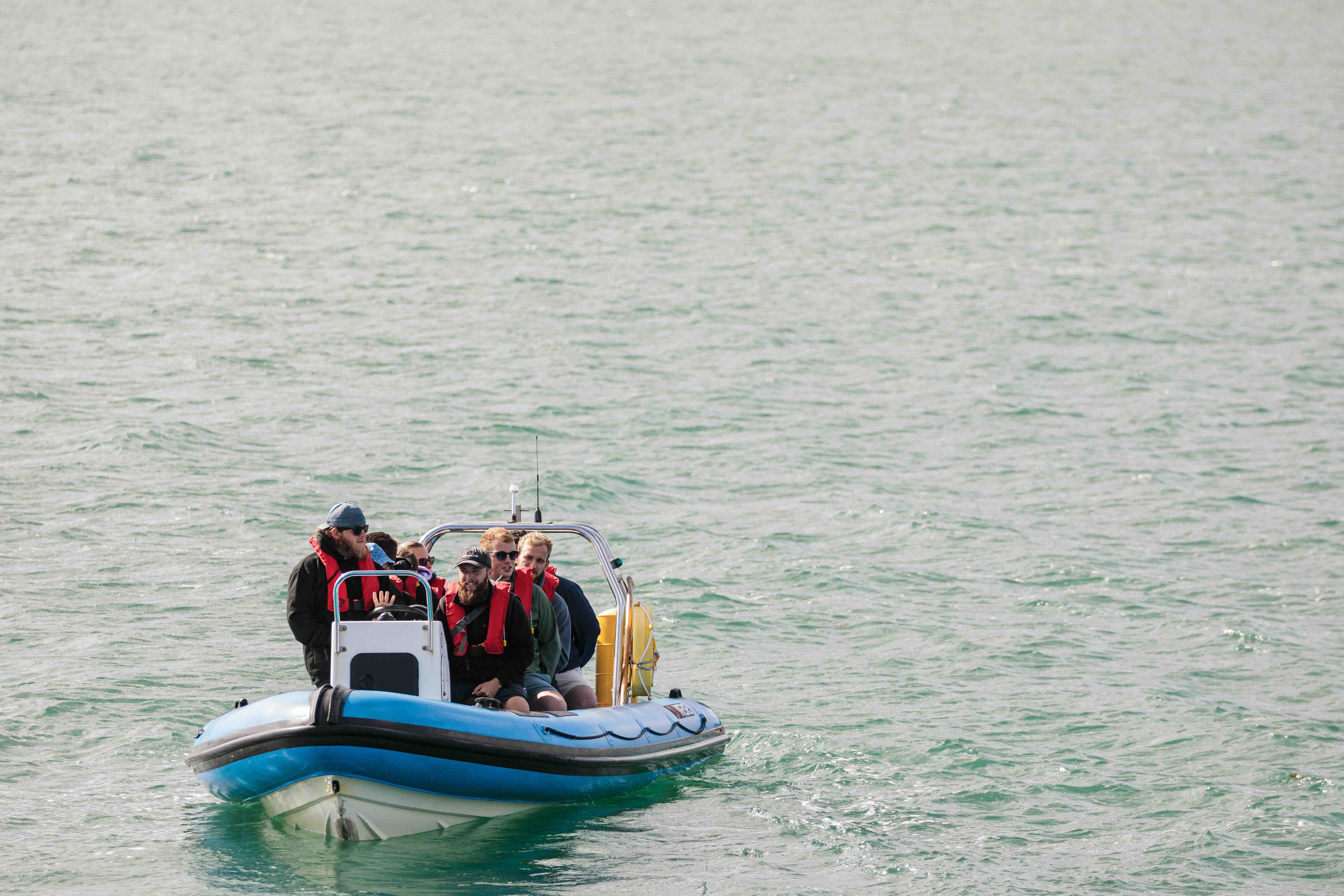 Group of six people wearing red life jackets on a small blue and white motorboat on calm water.