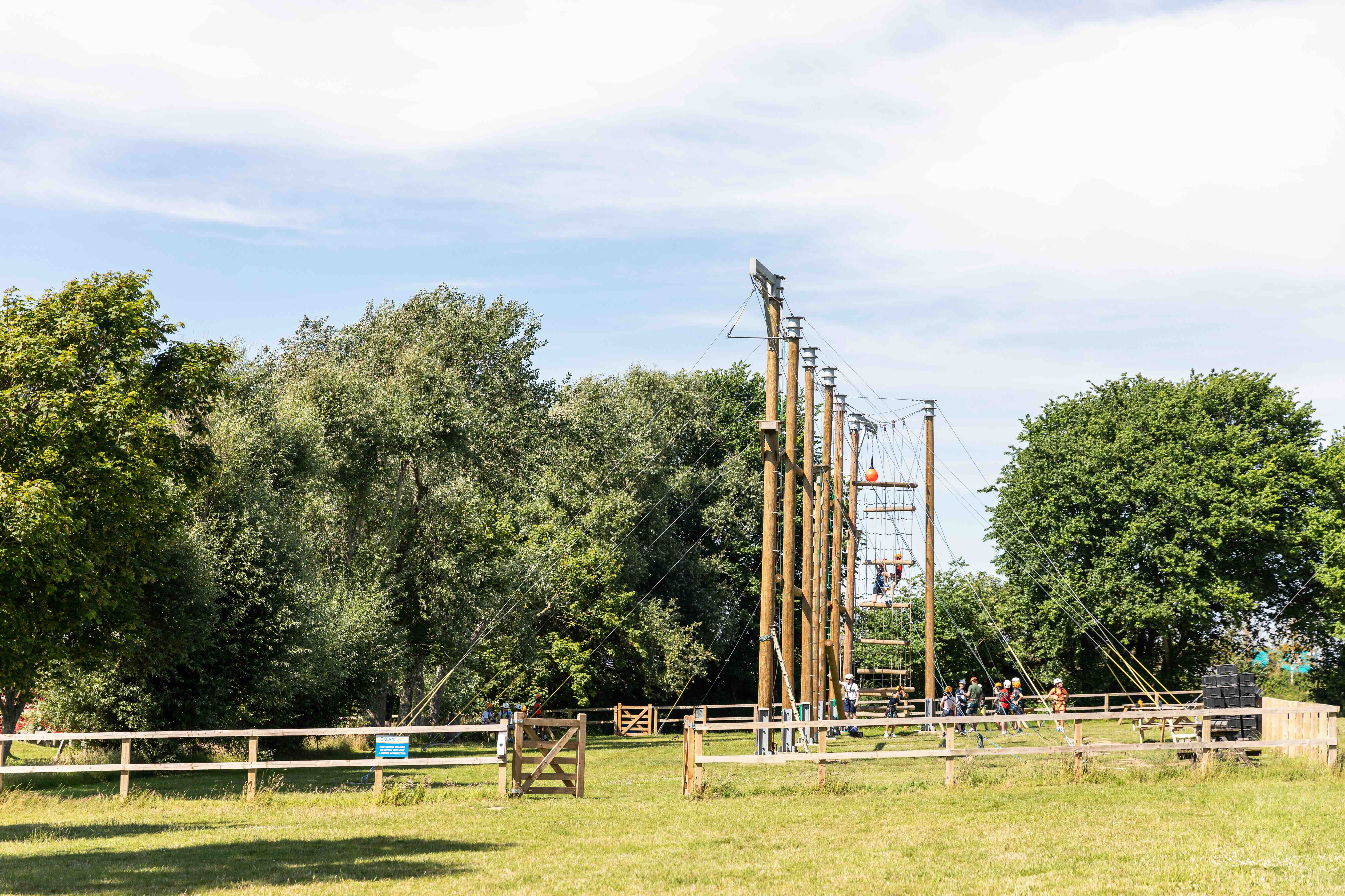Group of people wearing helmets preparing on ropes course with wooden poles in a grassy outdoor area surrounded by trees.
