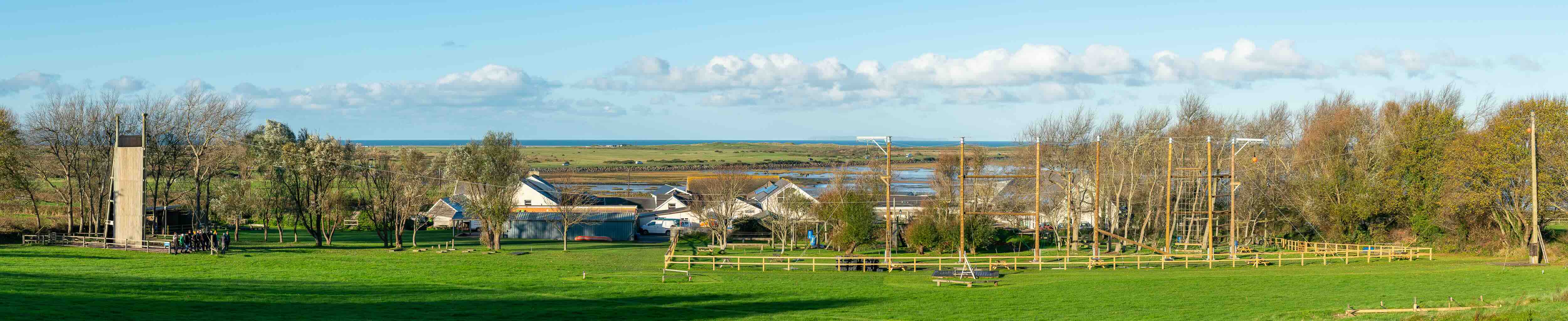Outdoor high ropes course with tall wooden climbing structures and green fields under a blue sky with scattered clouds.