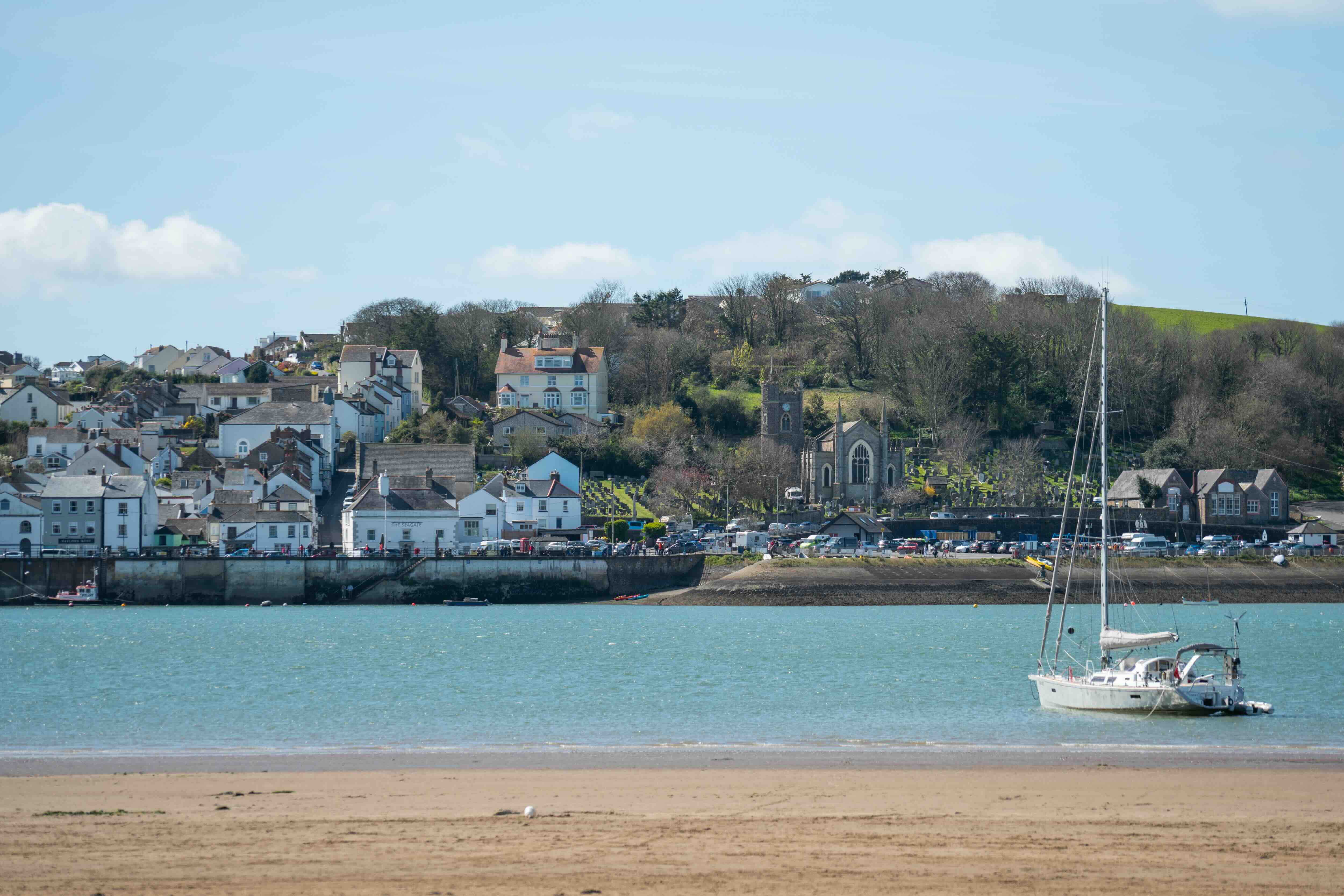 White sailboat anchored on calm water with a coastal town featuring houses and a church in the background under a blue sky.
