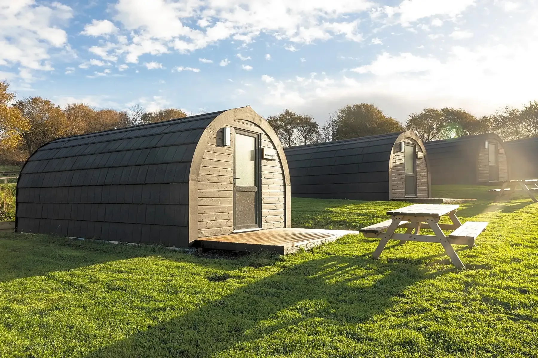 Three curved wooden glamping pods on green grass with picnic tables and trees under a partly cloudy sky during sunset.