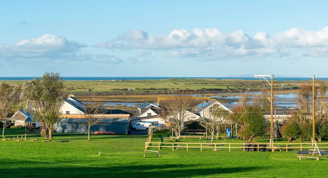 Rural coastal scene with green fields, houses, leafless trees, and the sea under a blue sky with scattered clouds.