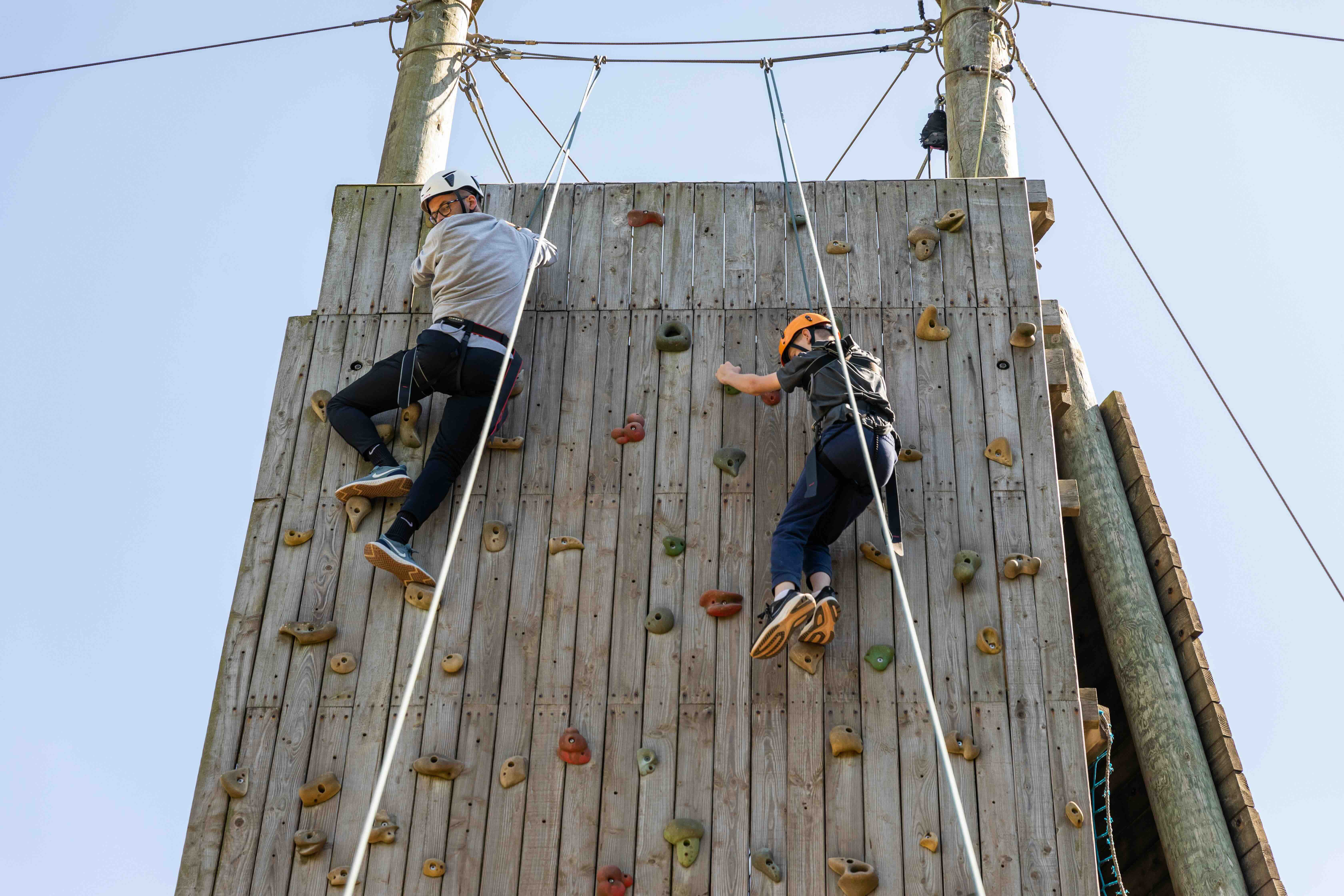Two people wearing helmets and harnesses climbing a wooden outdoor climbing wall with holds, under a clear blue sky.