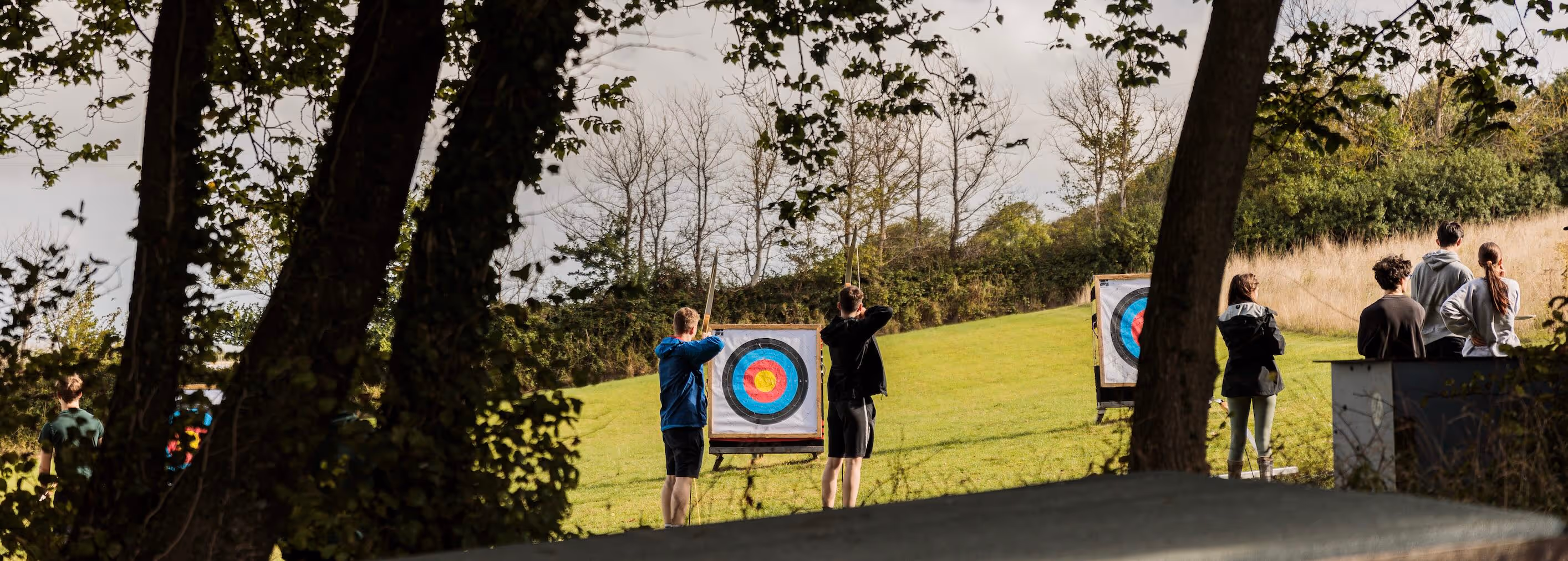 People practicing archery outdoors on a grassy field with trees and target boards.