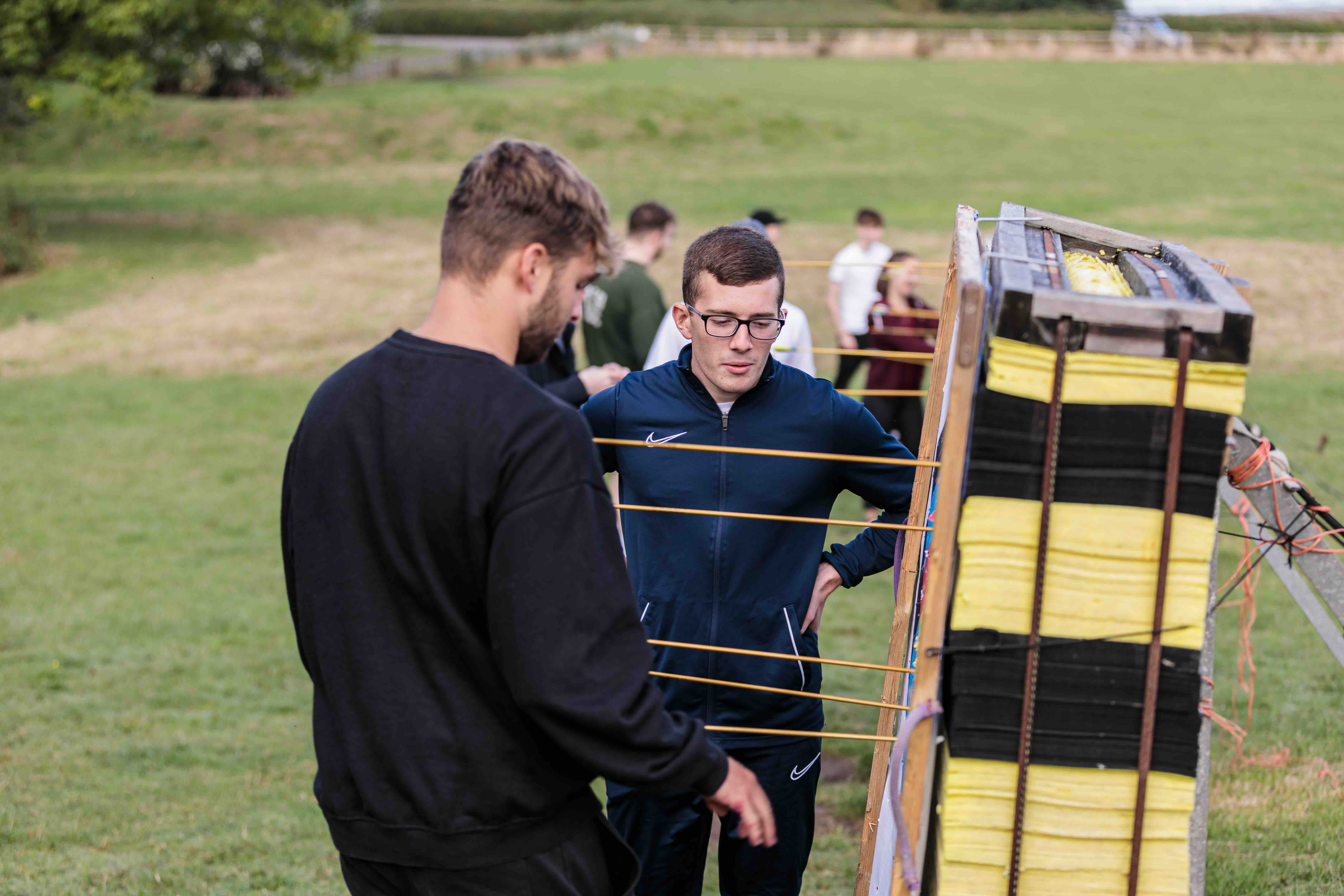 Two young men inspecting an archery target board with multiple arrows stuck in it on a grassy field.