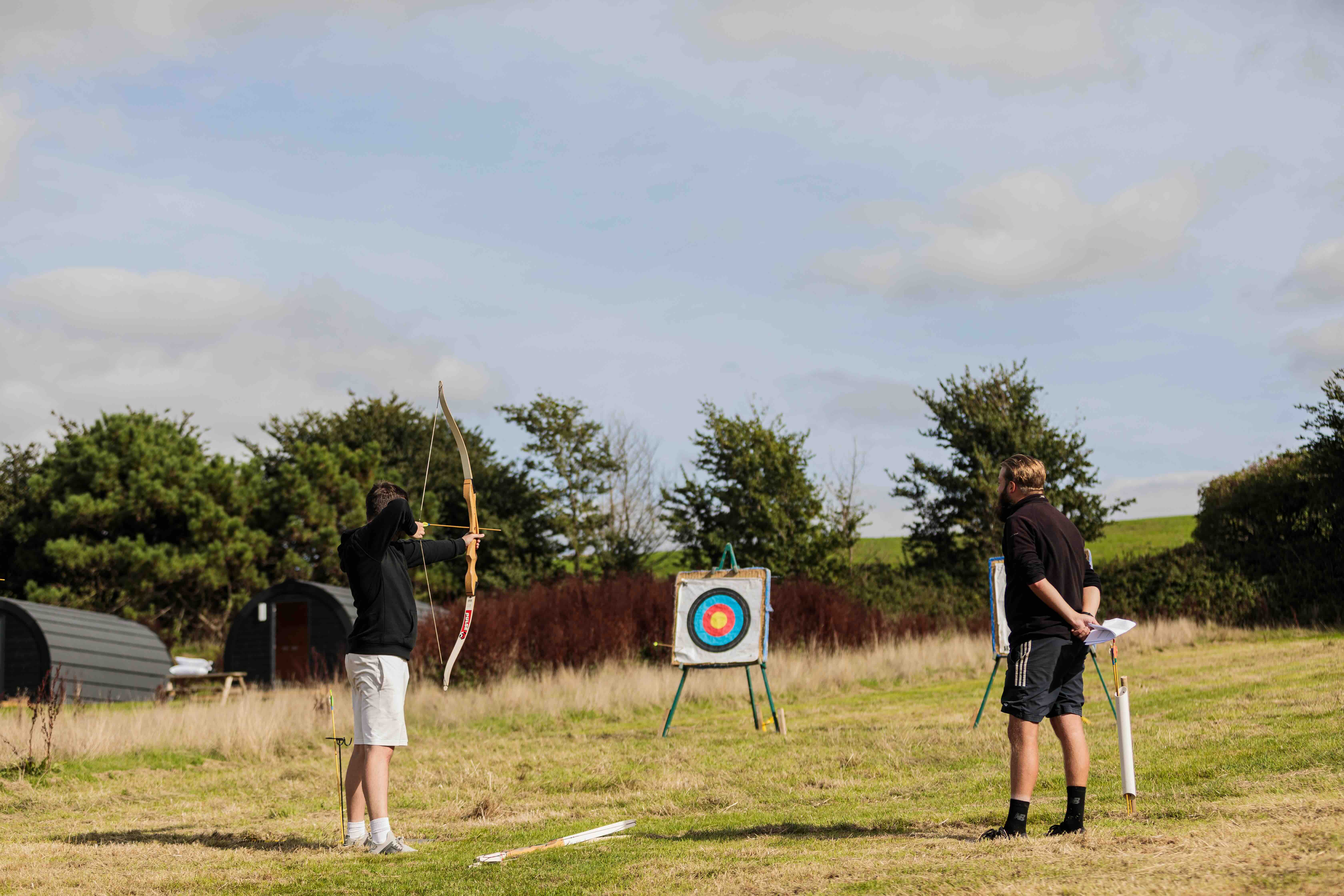 A young man aiming a bow towards a target while another man watches in an outdoor archery range.