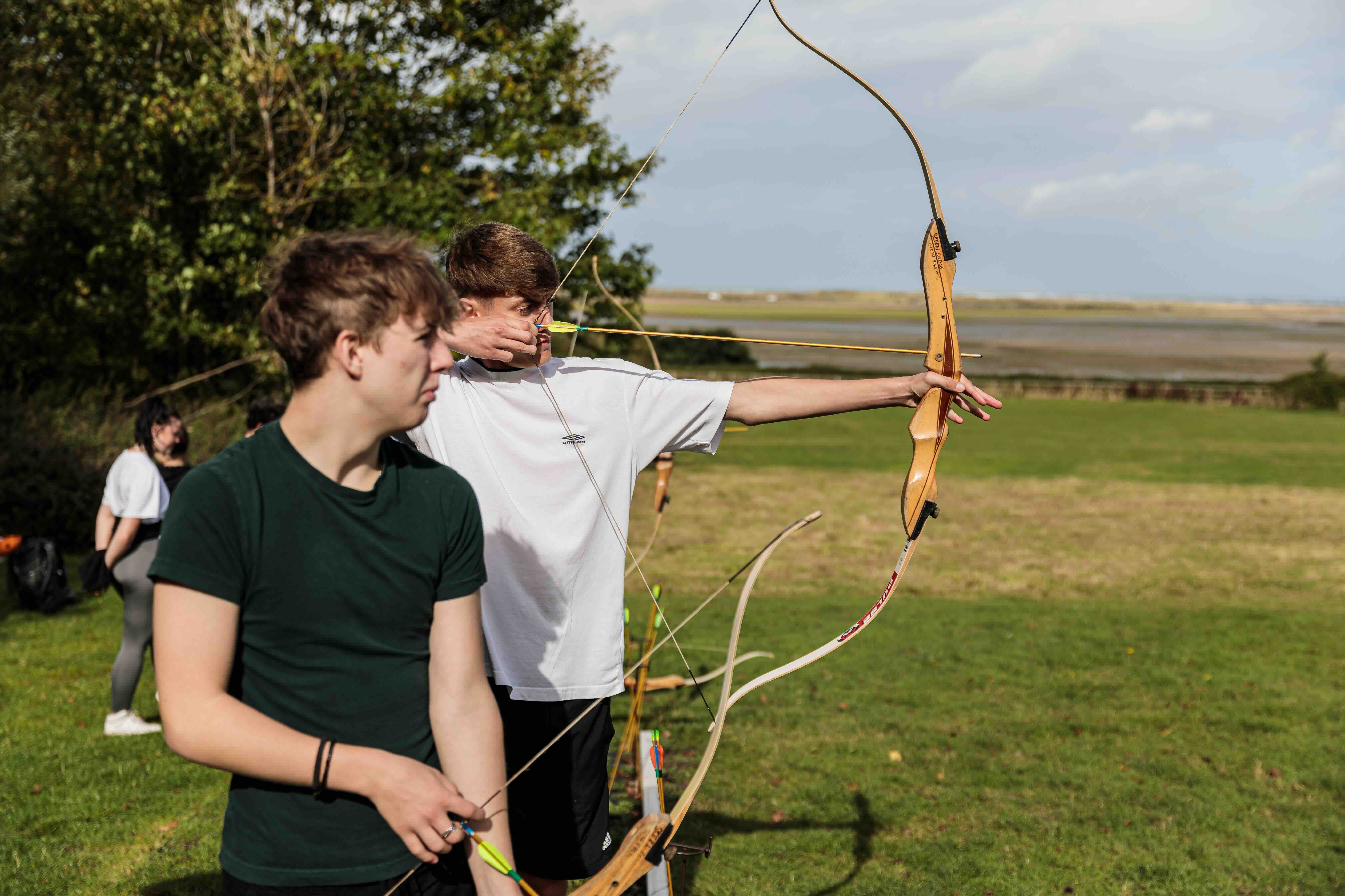 Two teenage boys outdoors practising archery, one aiming with a bow and arrow and the other holding a bow, with a grassy field and trees in the background.