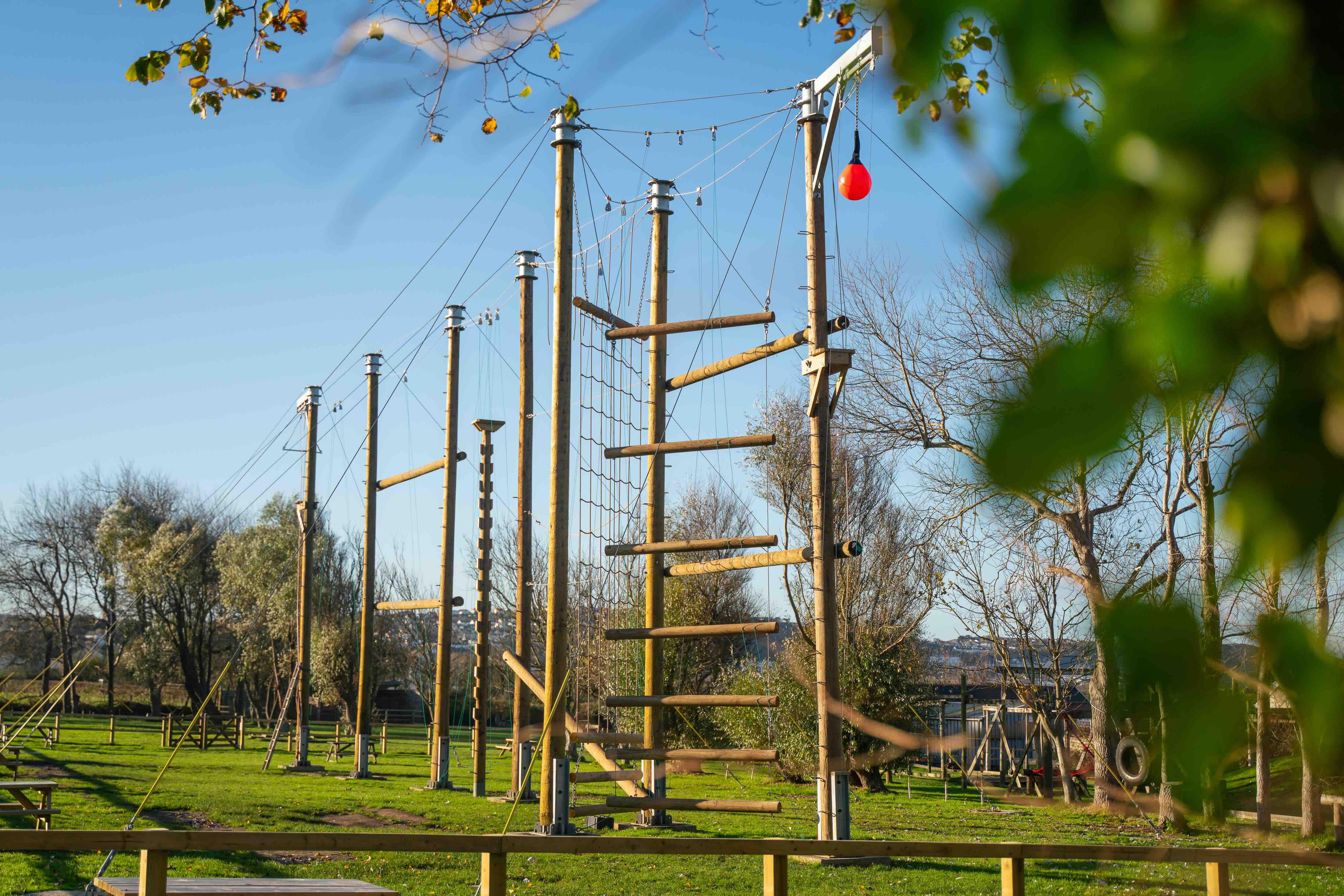 Outdoor high ropes assault course with wooden poles, climbing nets, and platforms set on a grassy field under a clear blue sky.