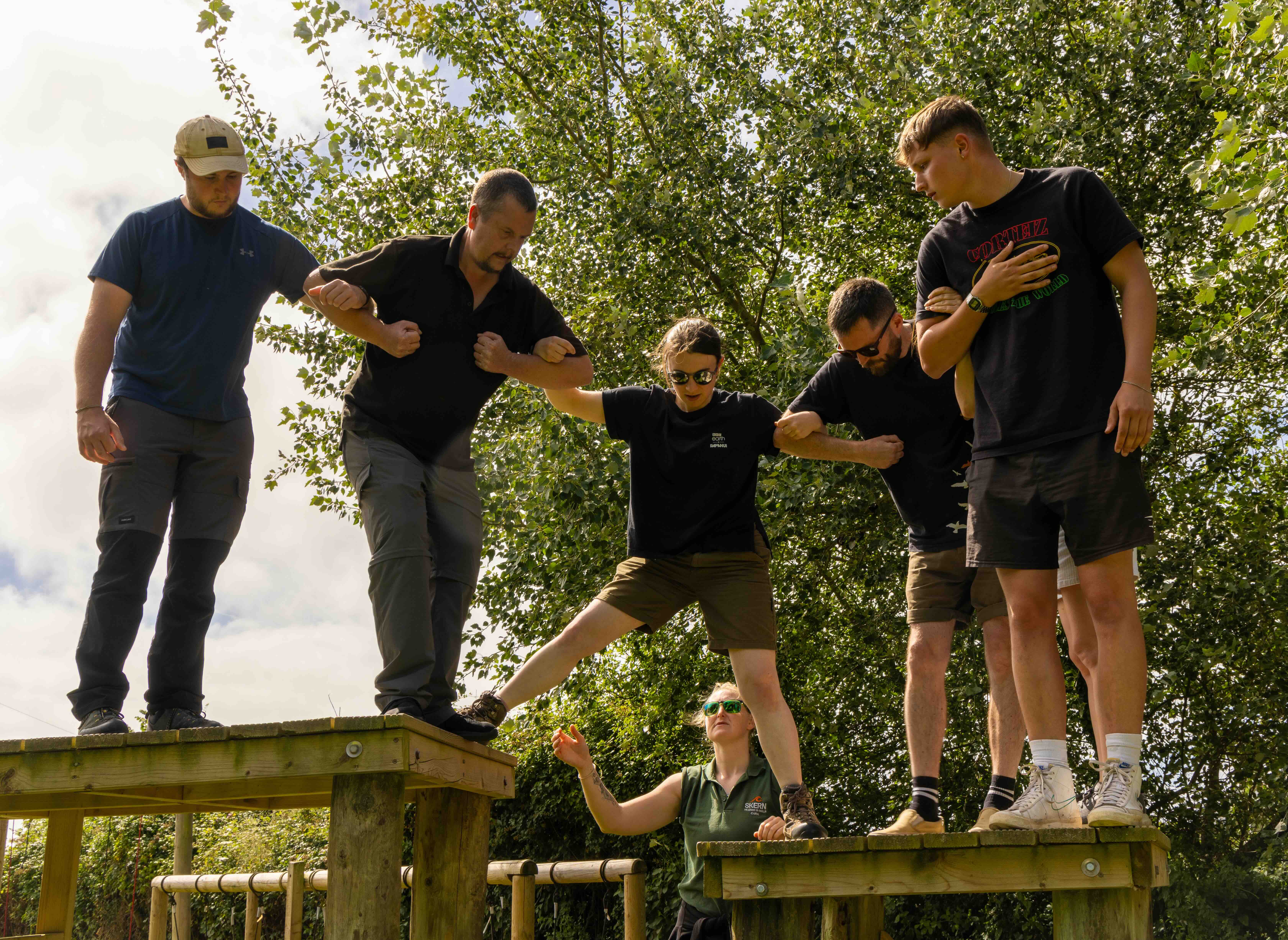 Group of five people holding arms to support a woman stepping across wooden platforms with an instructor guiding her below.