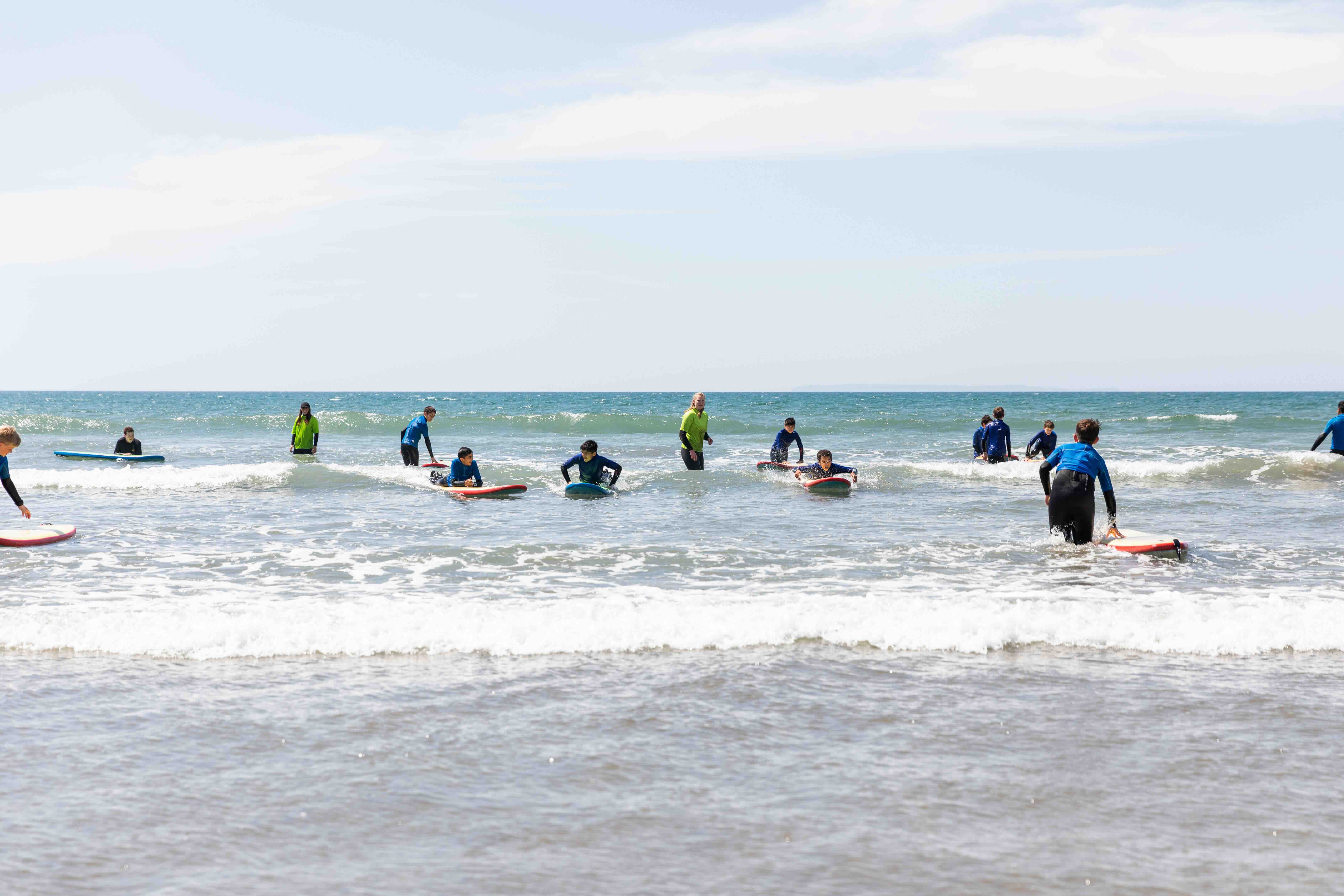 Group of children in wetsuits learning to surf with instructors in shallow ocean waves on a sunny day.