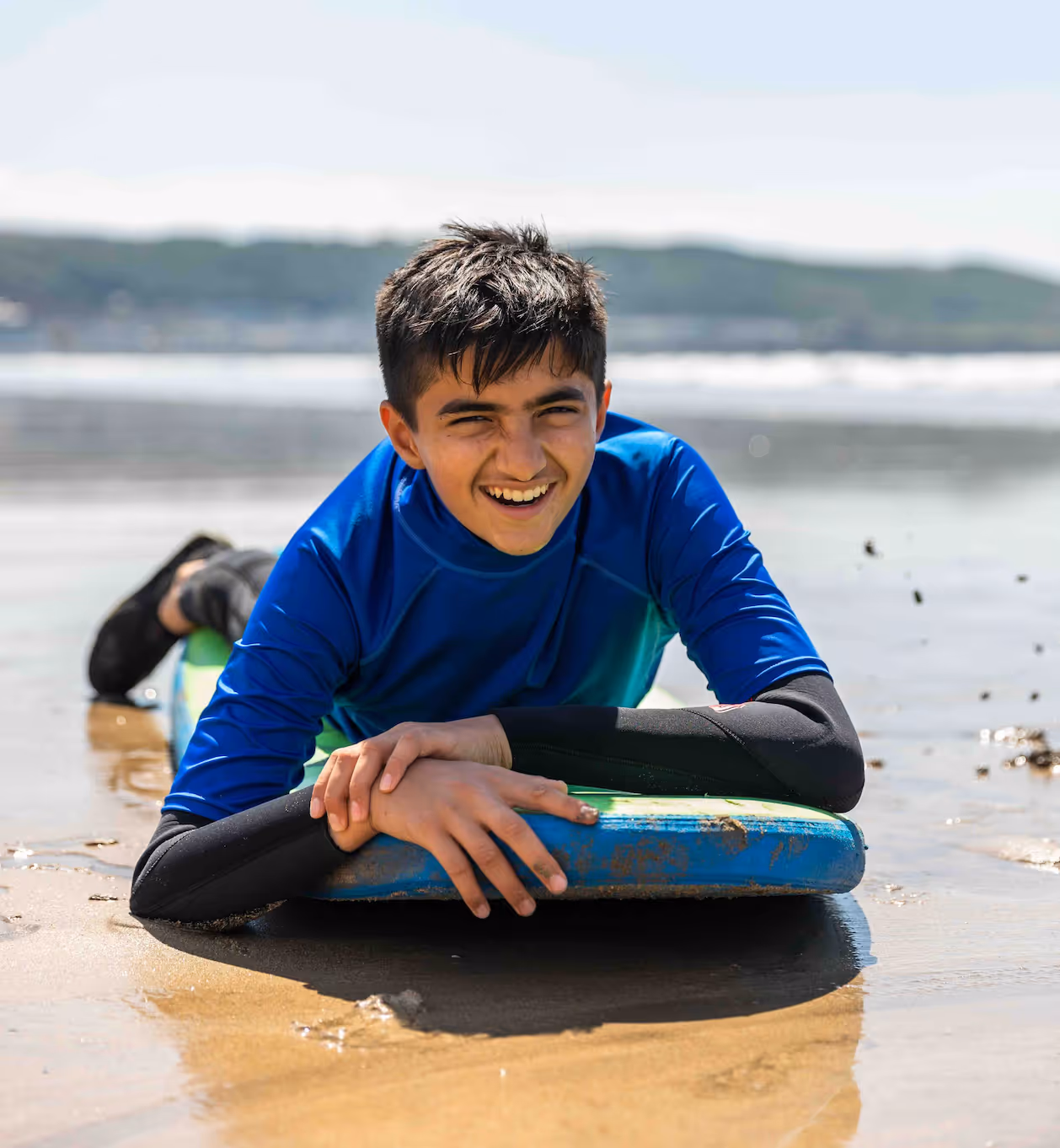 Smiling boy in blue swimwear lying on a bodyboard at the beach with water and hills in the background.