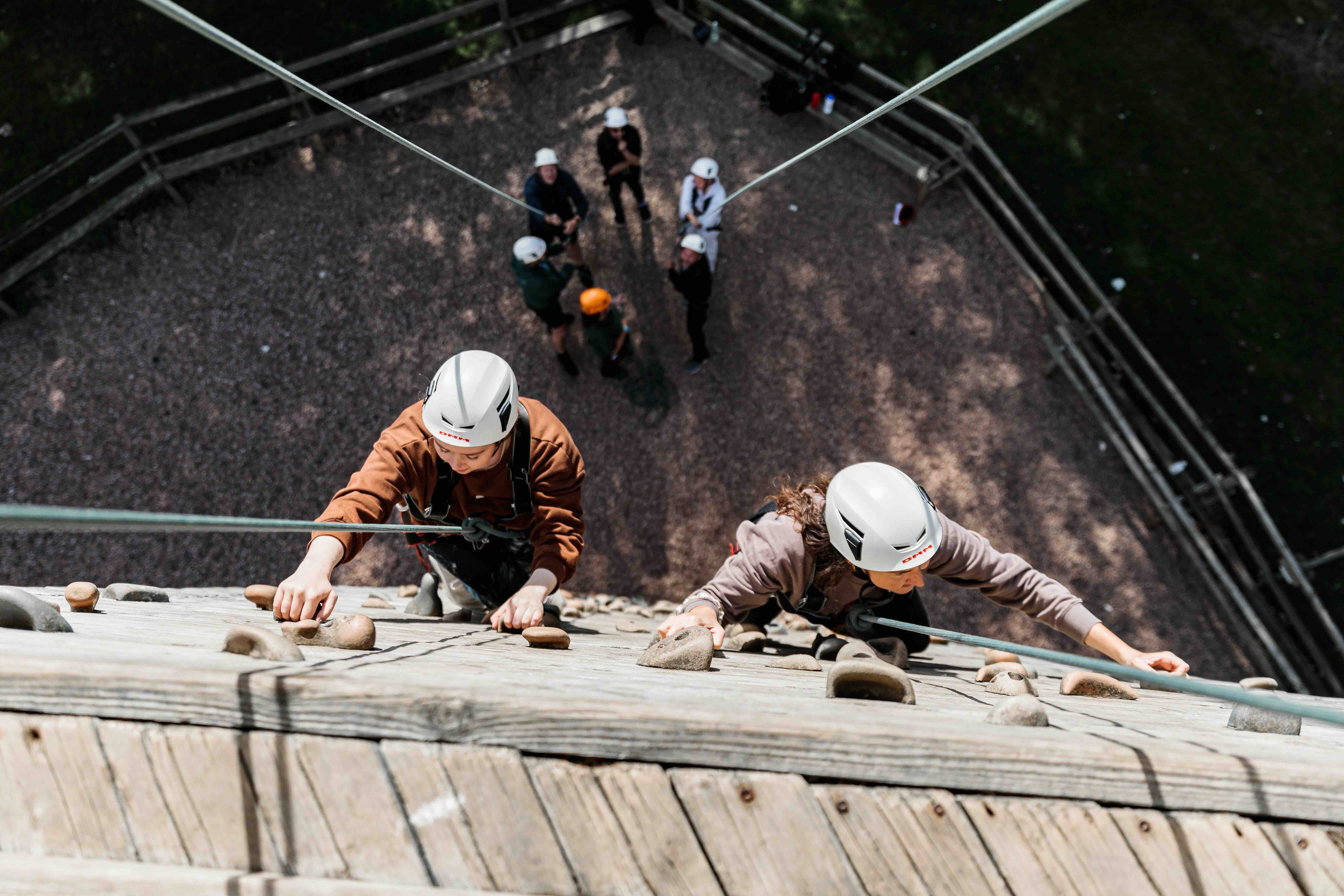 Two people wearing helmets and harnesses climbing a wooden climbing wall with handholds, viewed from above.