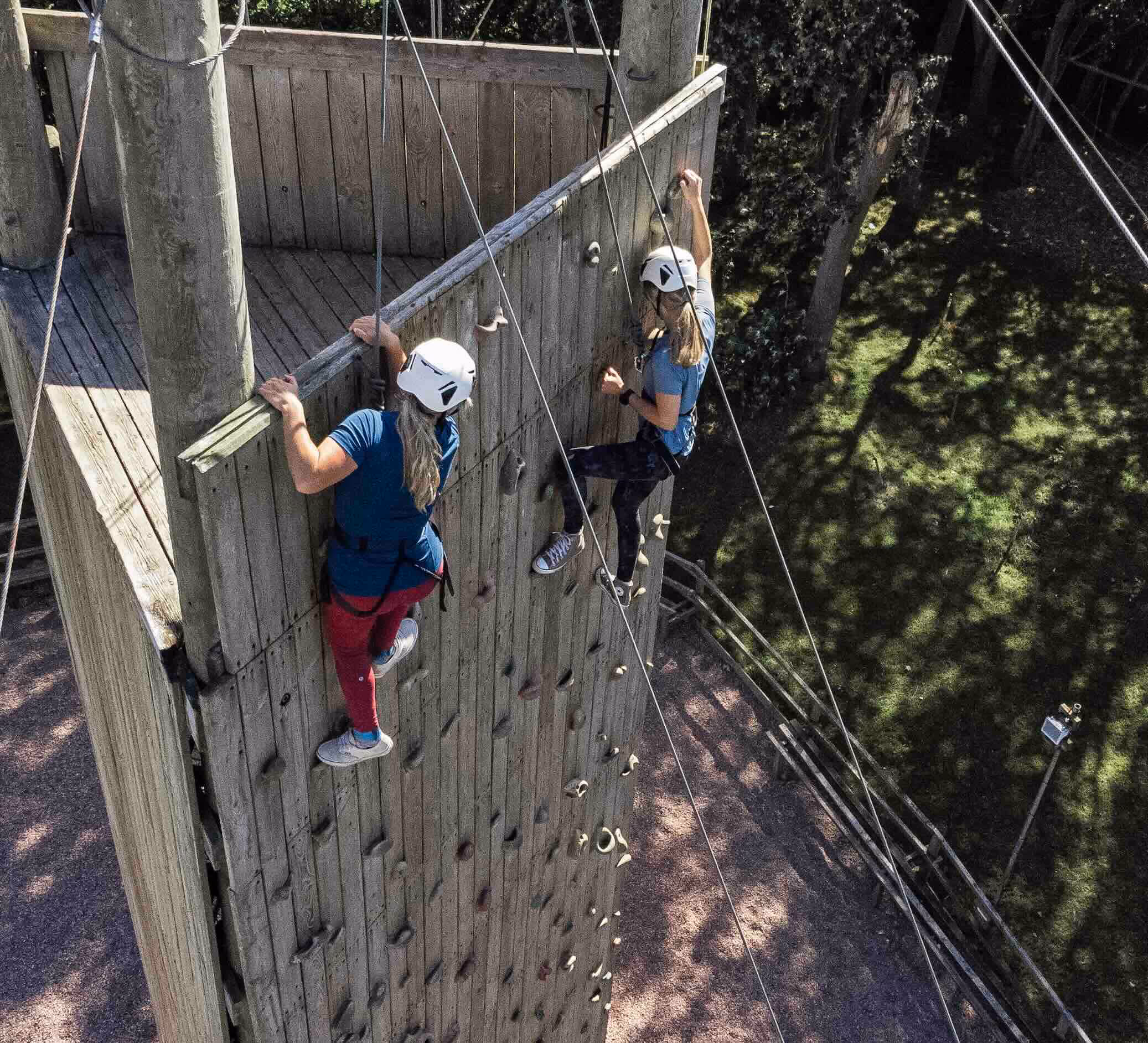 Two people wearing helmets and harnesses climbing a wooden rock climbing wall outdoors.