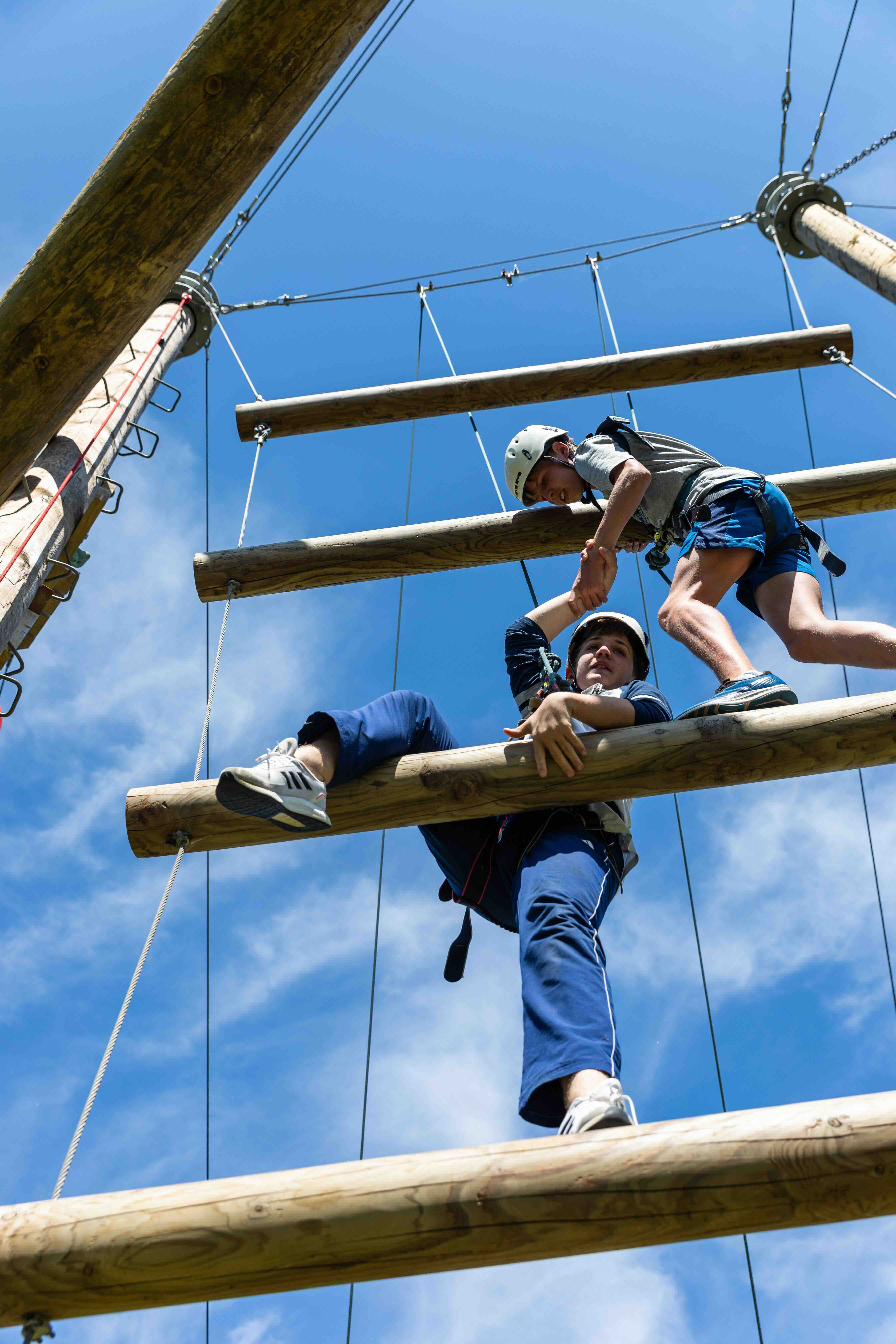 Two people wearing helmets and harnesses on a high ropes course helping each other climb wooden beams against a blue sky.