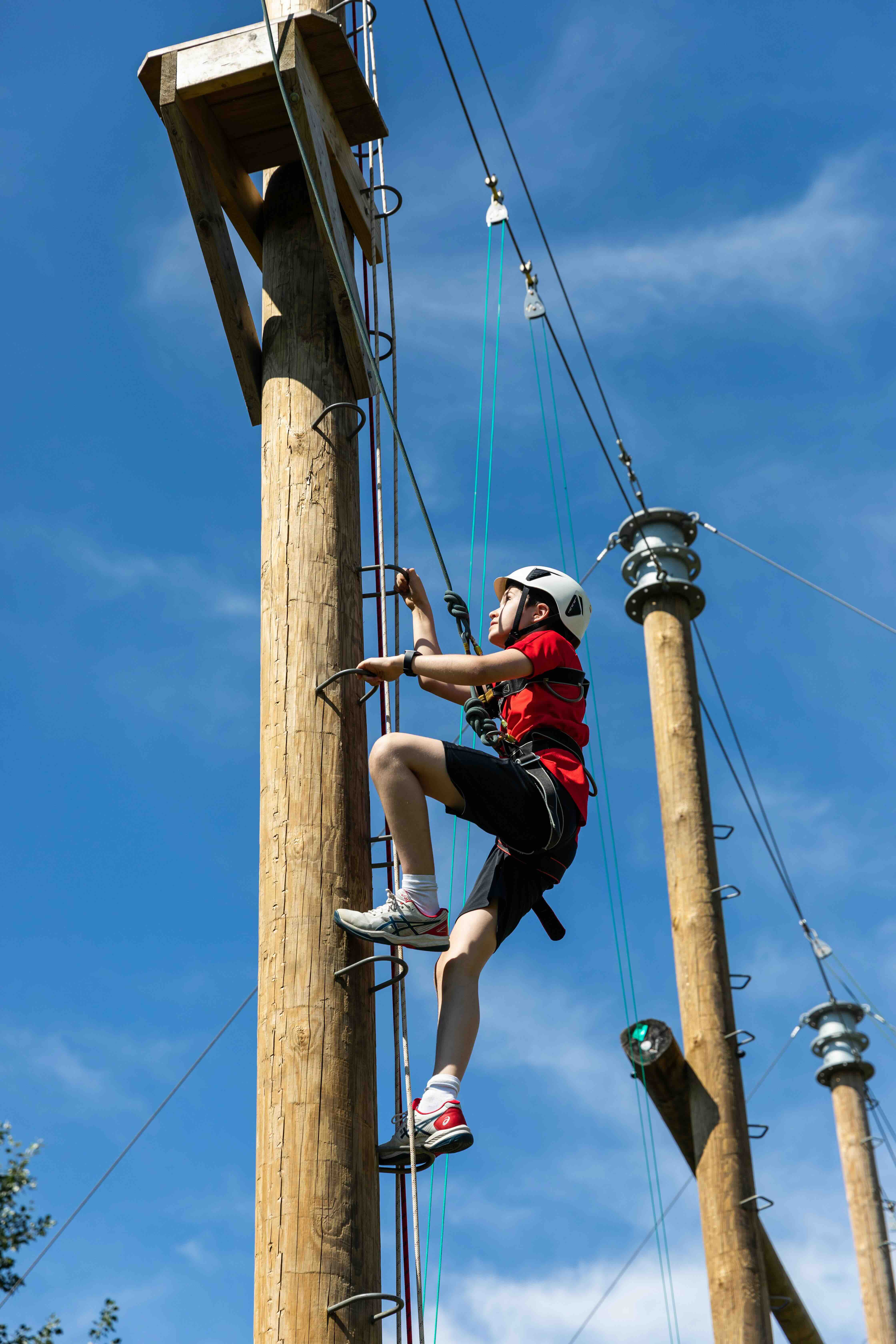 Child wearing helmet and safety harness climbing a wooden pole on a high ropes course against a blue sky.