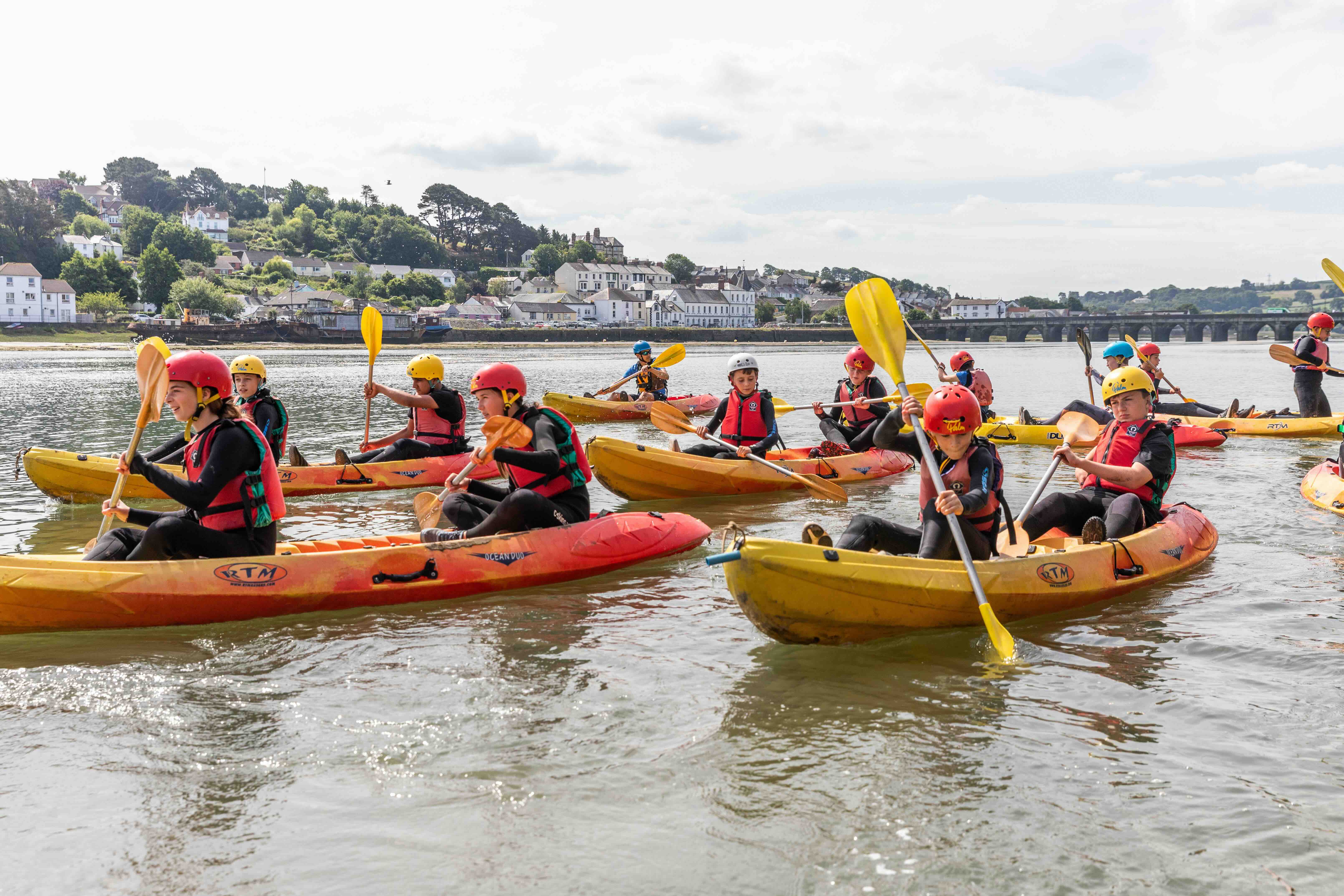 Group of children wearing helmets and life jackets paddling yellow and red kayaks on calm water near a riverside village.
