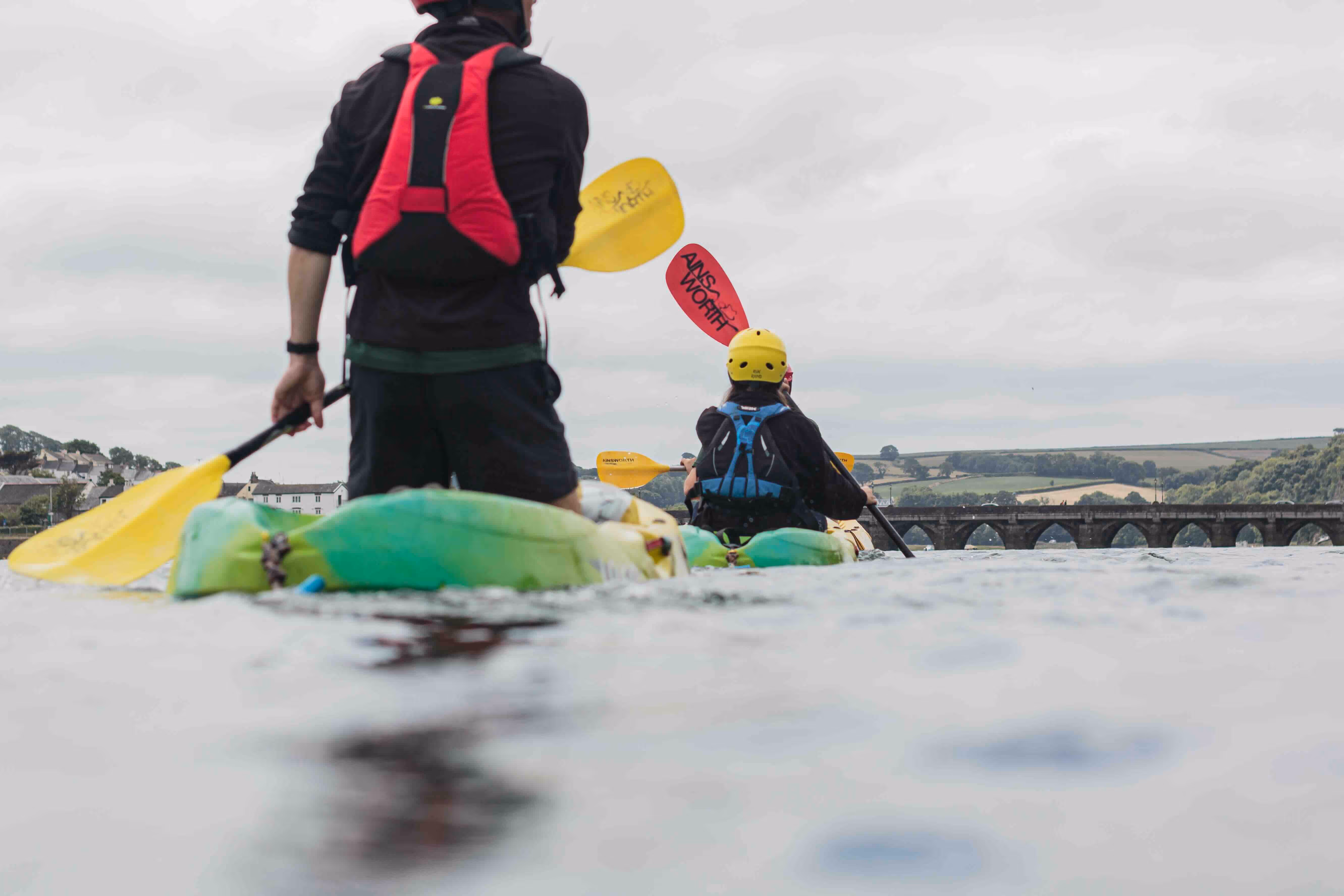Two people kayaking on a river heading towards a stone bridge with arched openings under a cloudy sky.